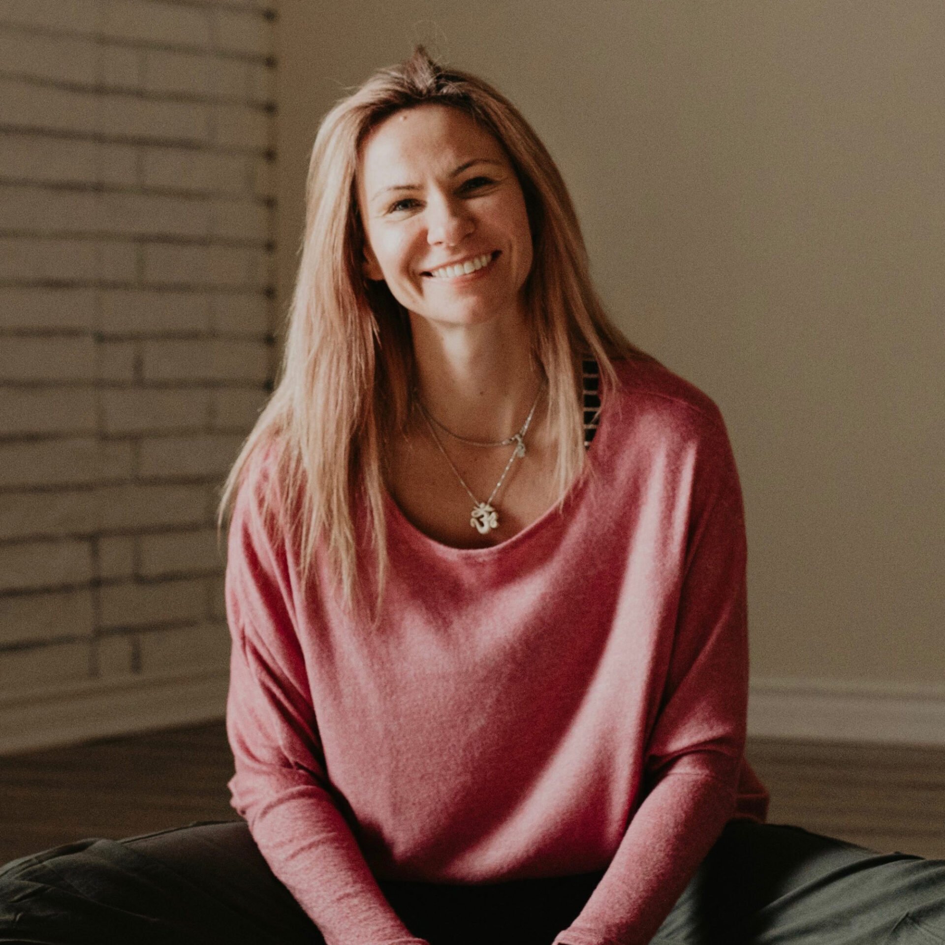 A woman with long, light brown hair, wearing a pink velvety long-sleeve shirt and layered necklaces, smiling while sitting on the floor indoors.