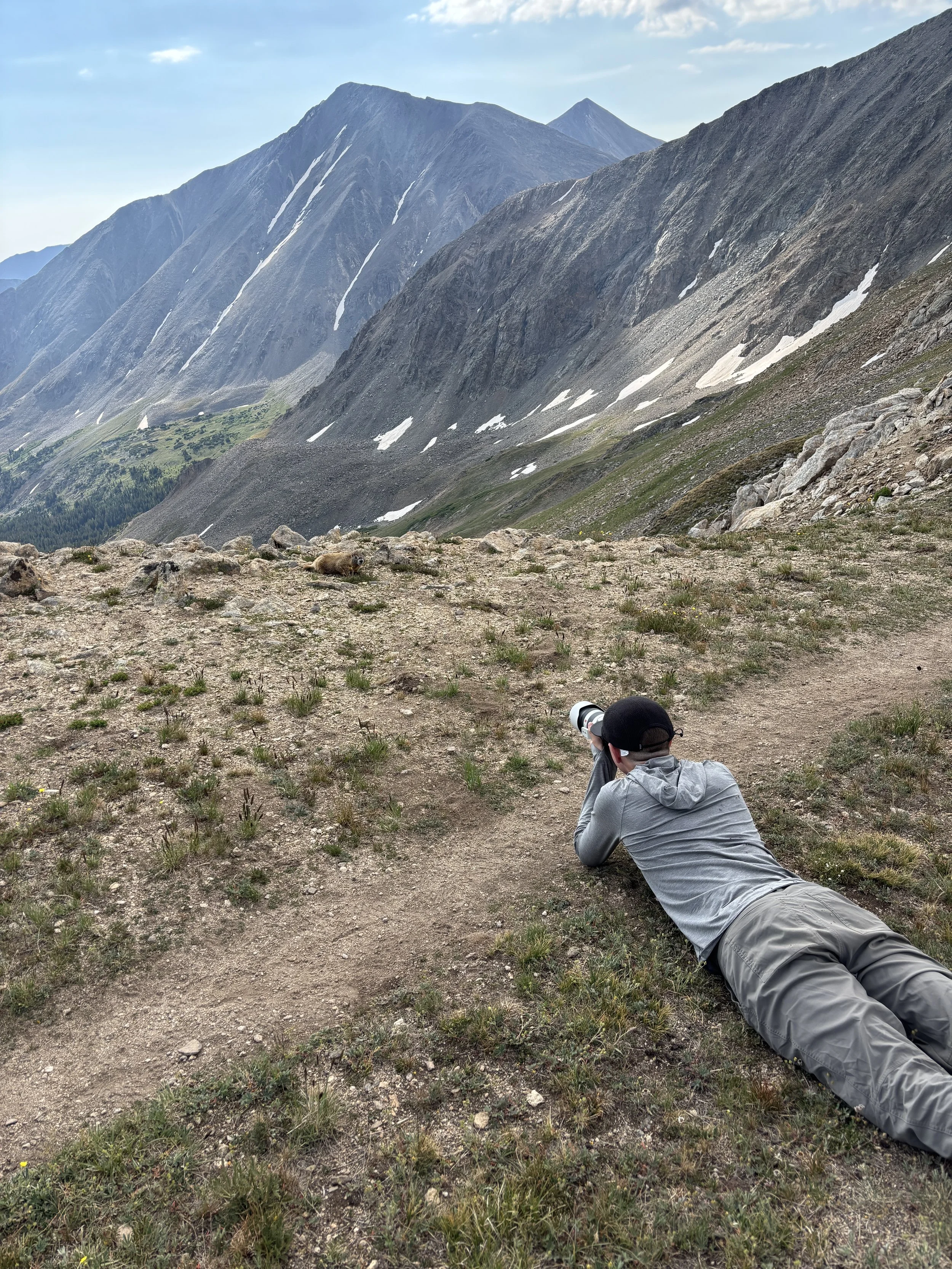 A person wearing gray outdoor clothing and a black cap lies on their stomach on a dirt trail, aiming a camera at a mountain landscape with rugged peaks and patches of snow. A marmot is lying on the ground in the distance near the rocky terrain.