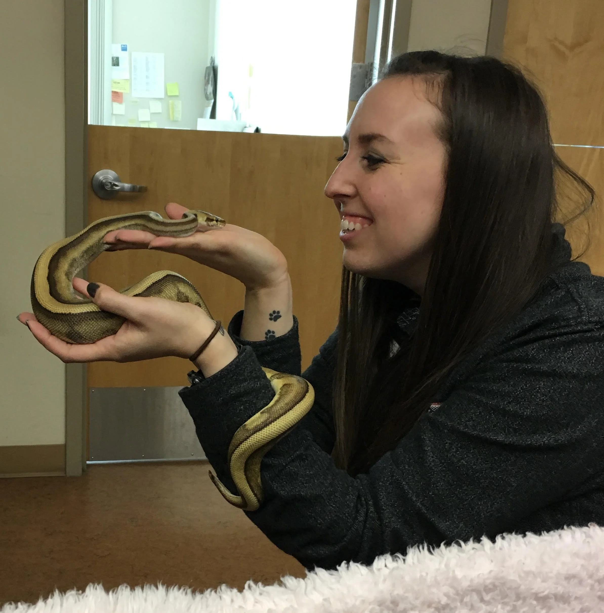 A woman holding a yellow and brown snake indoors, smiling and looking at the snake.