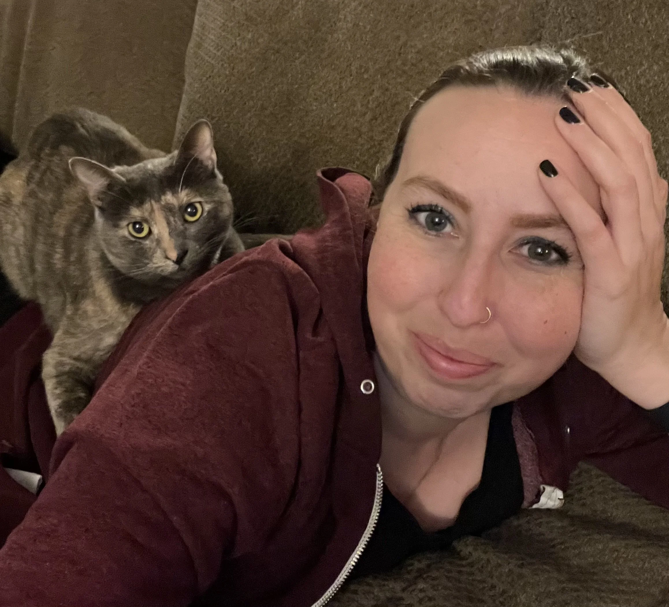 A woman with blue eyes and a nose ring lying on a couch, smiling, with a gray tabby cat on her back.