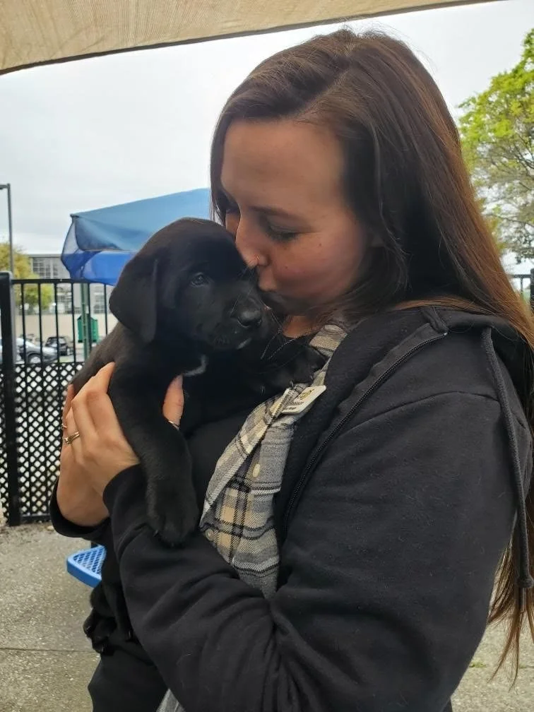 A woman with long brown hair holding and kissing a black puppy outdoors.