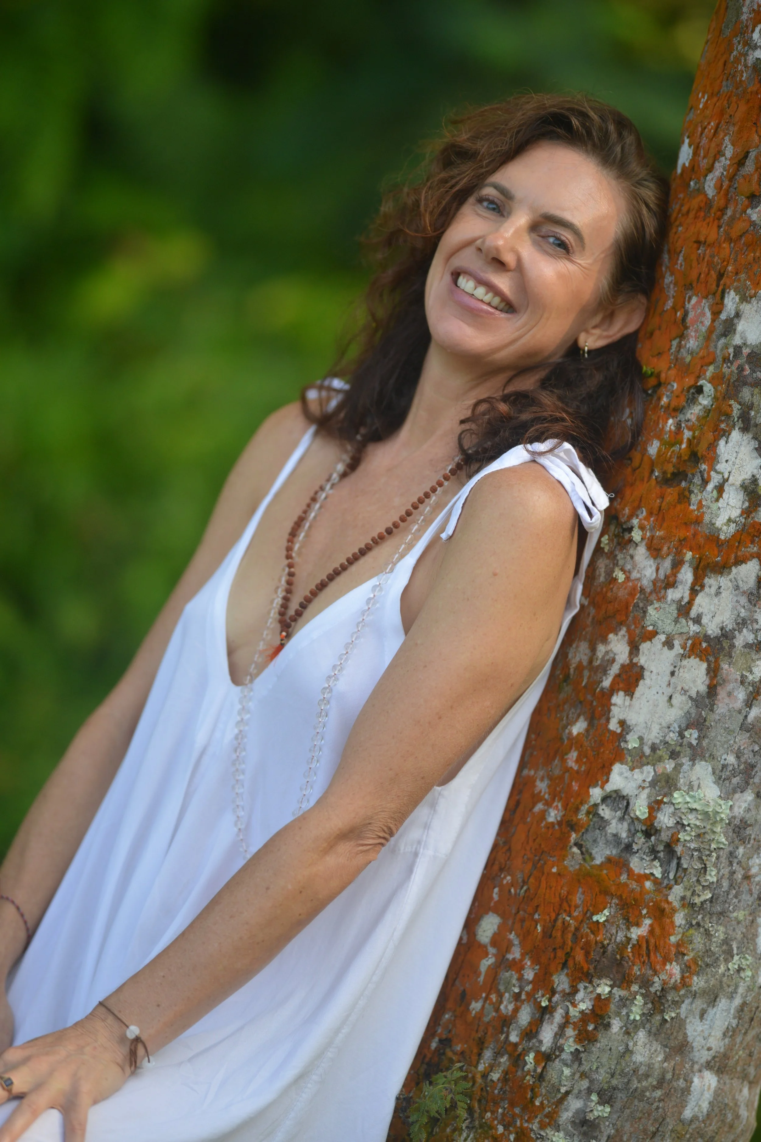A smiling woman with curly brown hair leaning against a tree outdoors, wearing a white sleeveless dress and beaded necklaces.