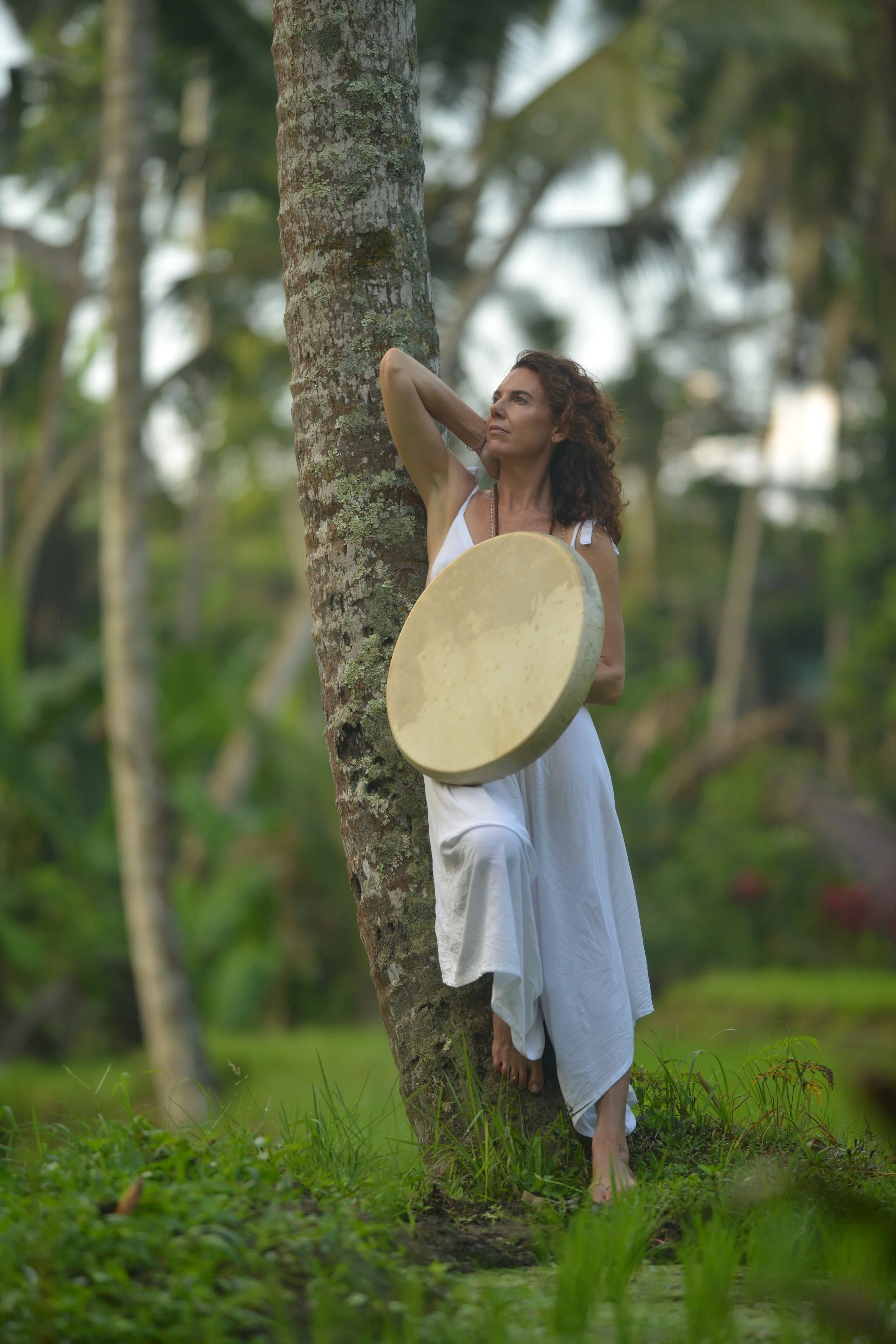 A woman in a white dress standing outdoors in a lush green area, leaning against a palm tree, holding a large round musical instrument, with her arm raised behind her head, gazing thoughtfully into the distance.