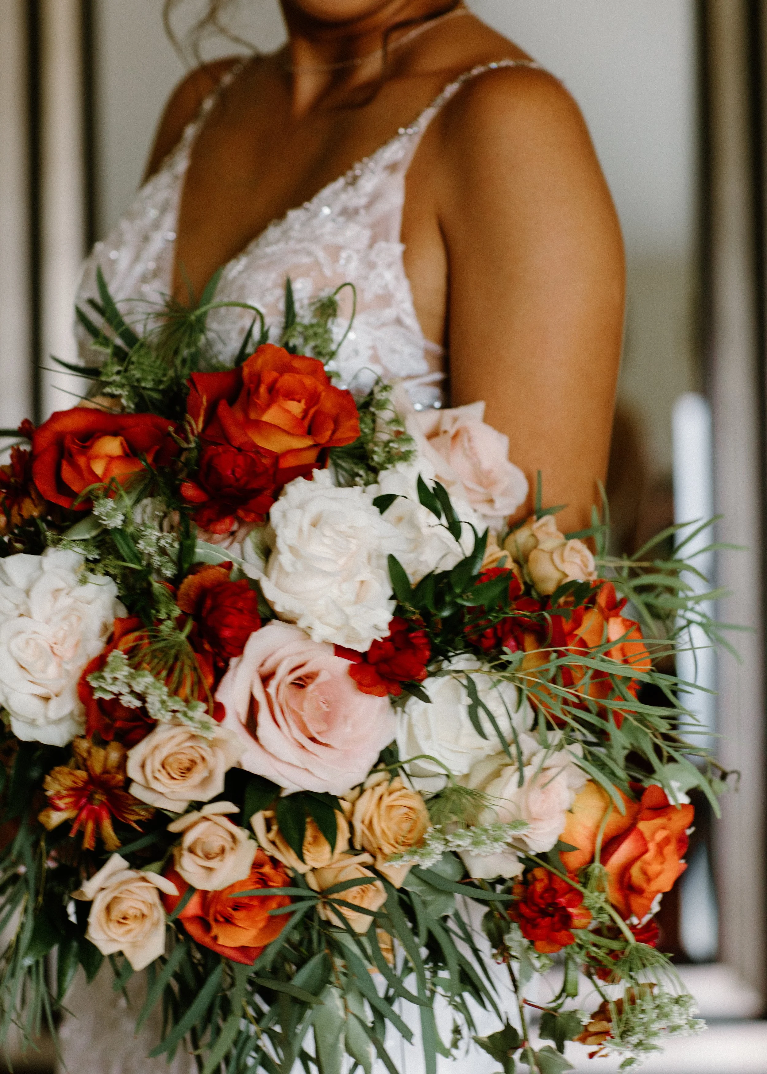 Close-up of a bride holding a large bouquet of orange, pink, and white roses, with green foliage, wearing a lace wedding dress with thin straps.