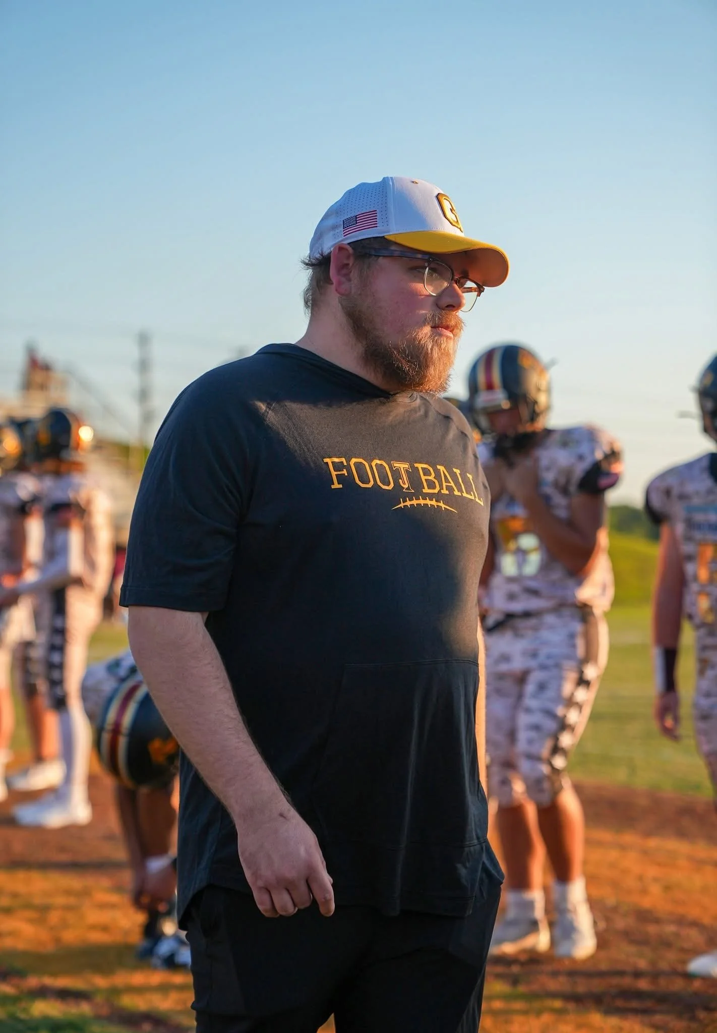 A man with a beard wearing a white hat, glasses, and a black T-shirt that says 'FOOTBALL' stands on a sports field during sunset, with football players in helmets and uniforms in the background.