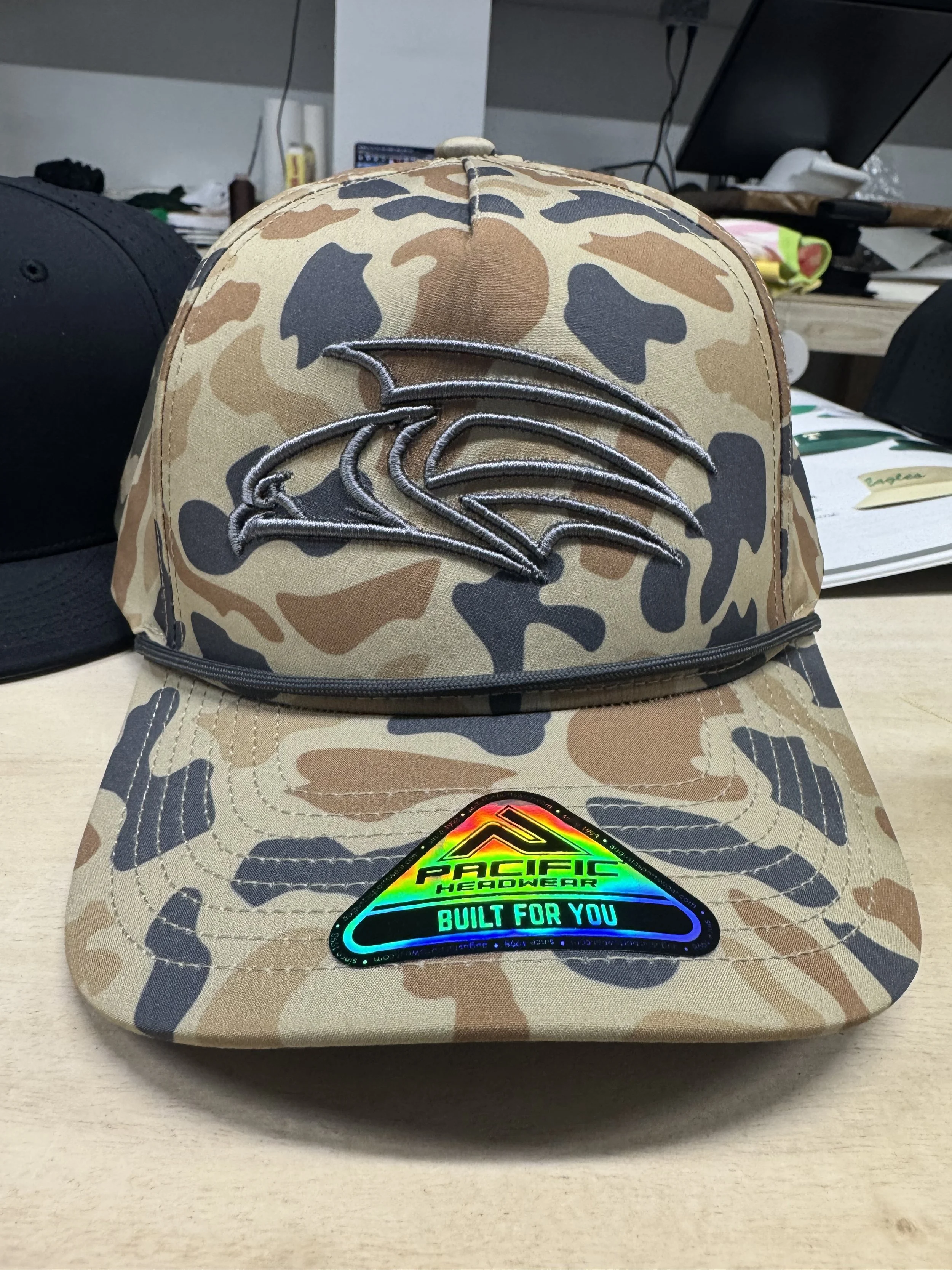 Camouflage baseball cap with a silver embroidered eagle logo on the front and a colorful holographic sticker on the brim.