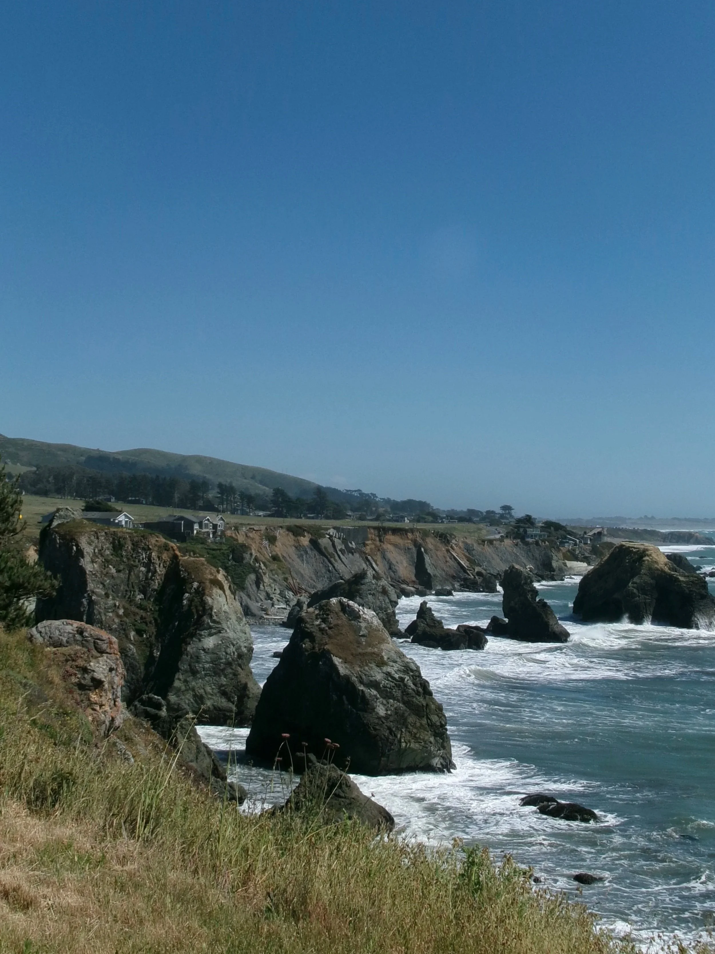 Coastal landscape with rocky cliffs, ocean waves, green grassy foreground, houses along the coast, and hills in the background under a clear blue sky.