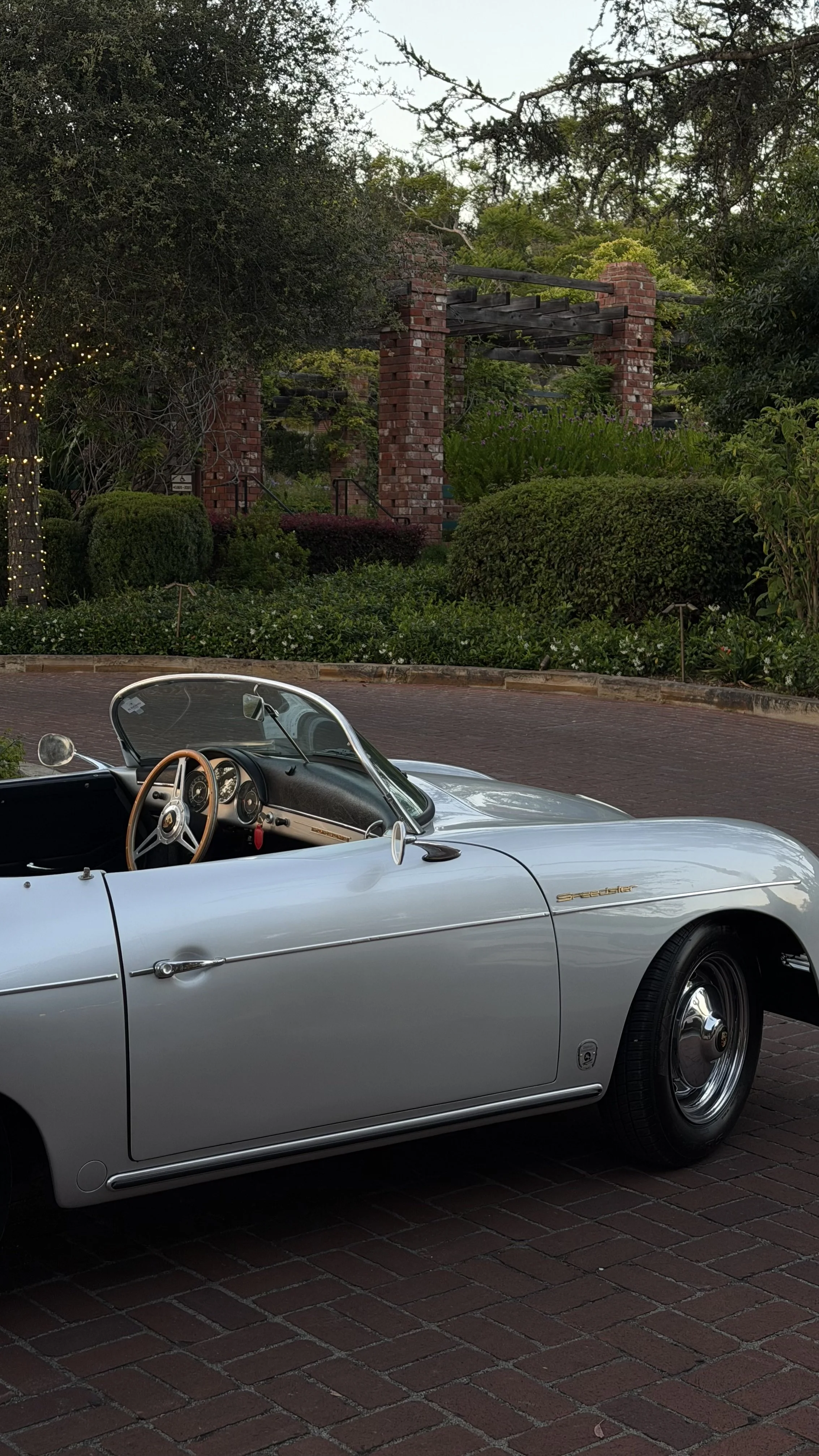A classic silver Porsche Speedster convertible parked on a brick street with greenery and a brick structure in the background.