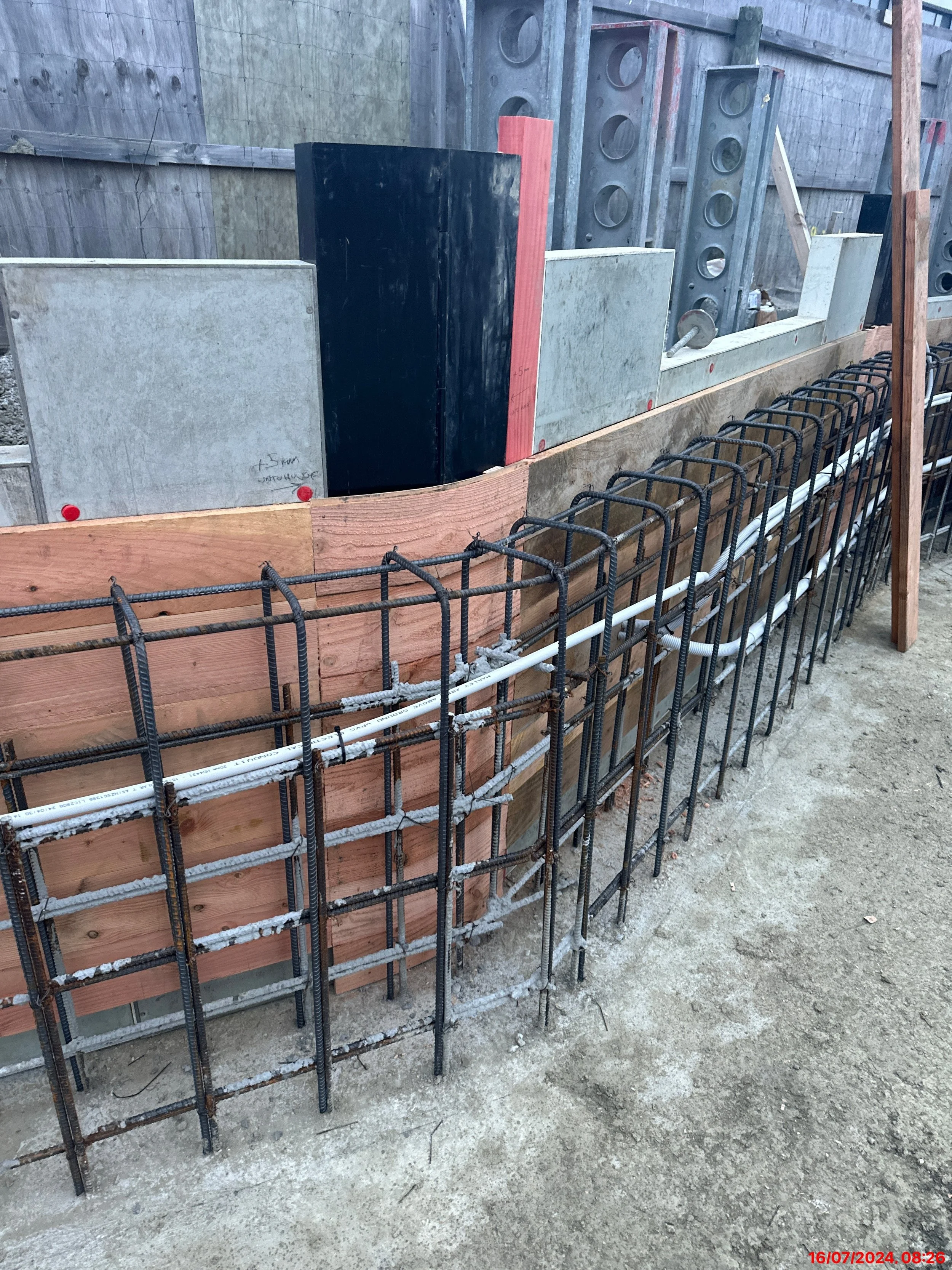 Construction site showing reinforced concrete foundation walls, wood formwork, rebar cages, and embedded utility pipes.