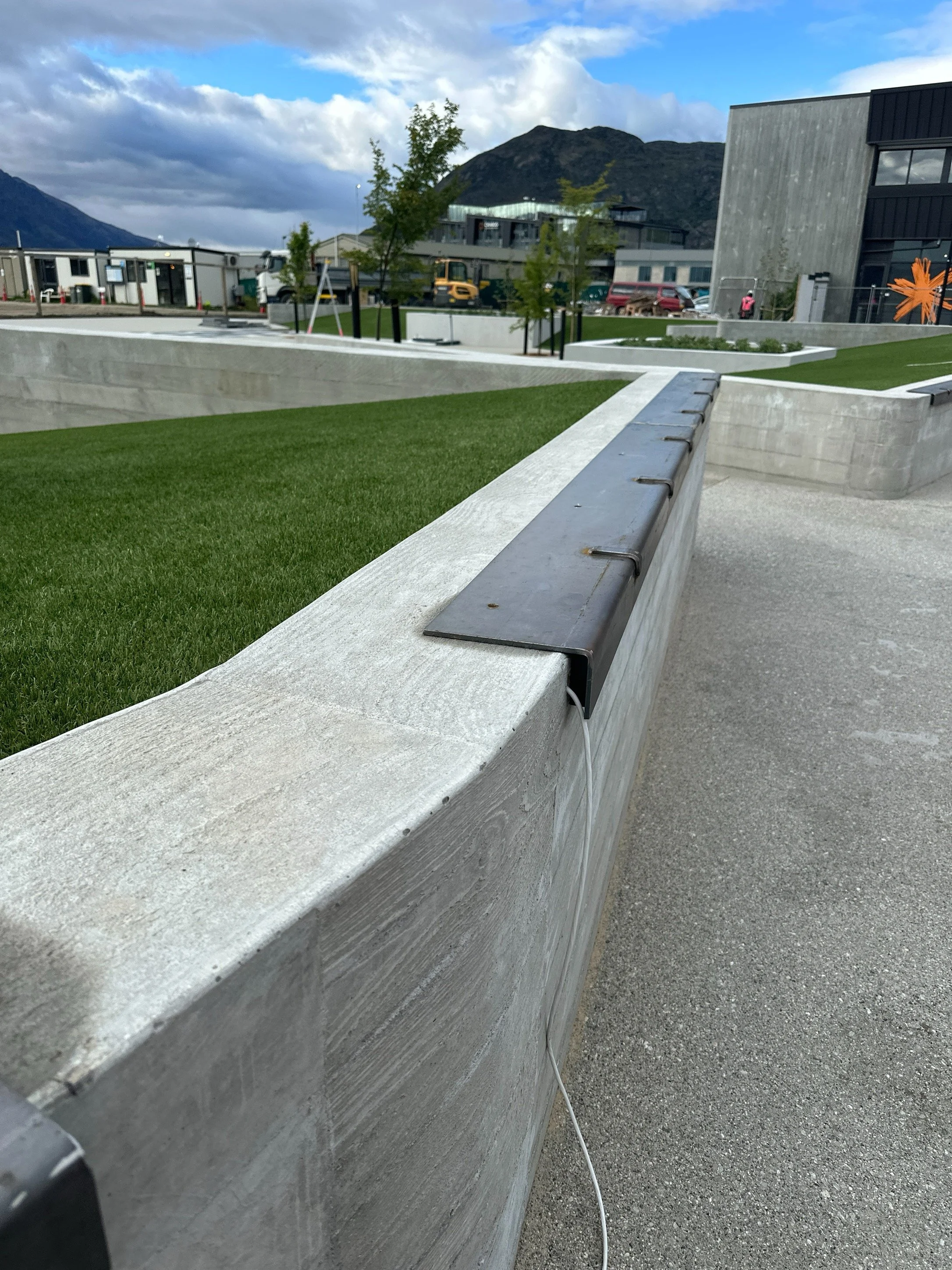 Modern concrete courtyard with landscaped lawn and buildings against rugged mountains under a cloudy blue sky
