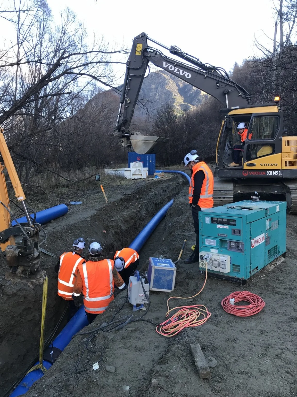 Construction crew installs a blue pipeline in a trench beside a Volvo excavator in a mountainous wooded area.