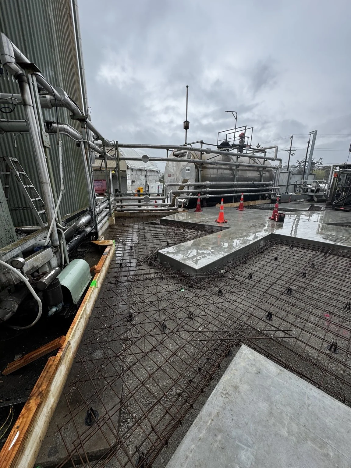Industrial rooftop construction site with exposed rebar, concrete forms, pipes, tanks, safety cones, and overcast skies