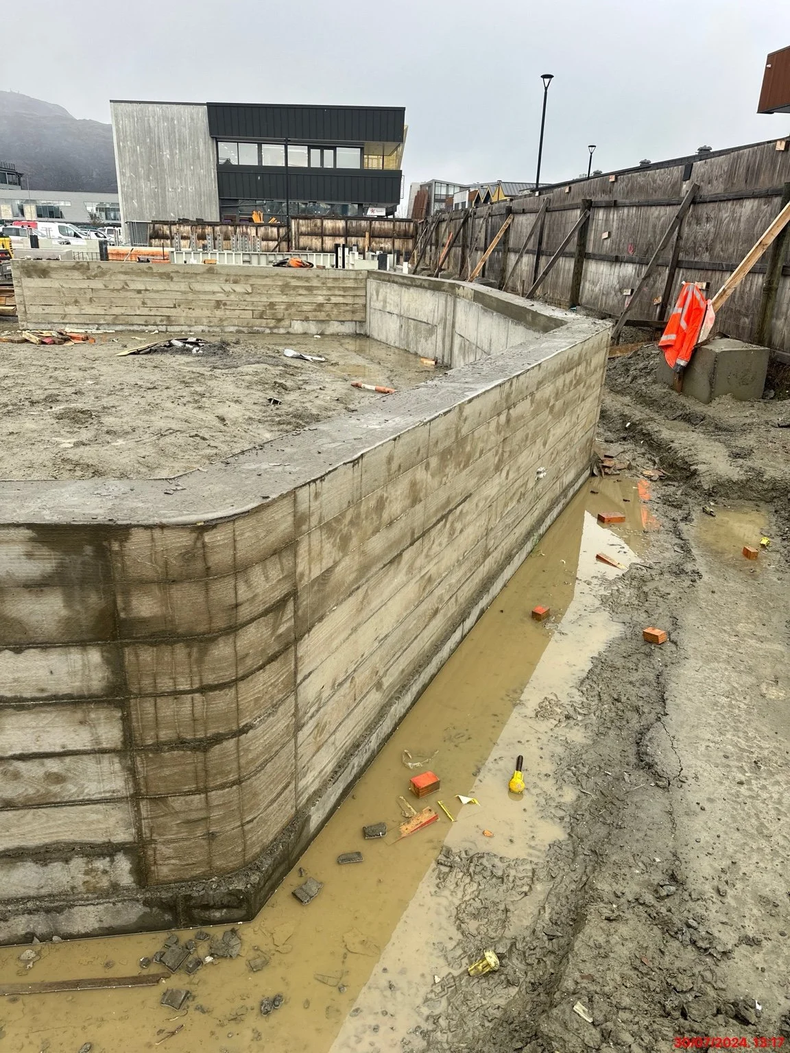 Construction site showing newly poured concrete foundation walls, muddy standing water, temporary wooden fencing, and industrial buildings with mountains in the background.
