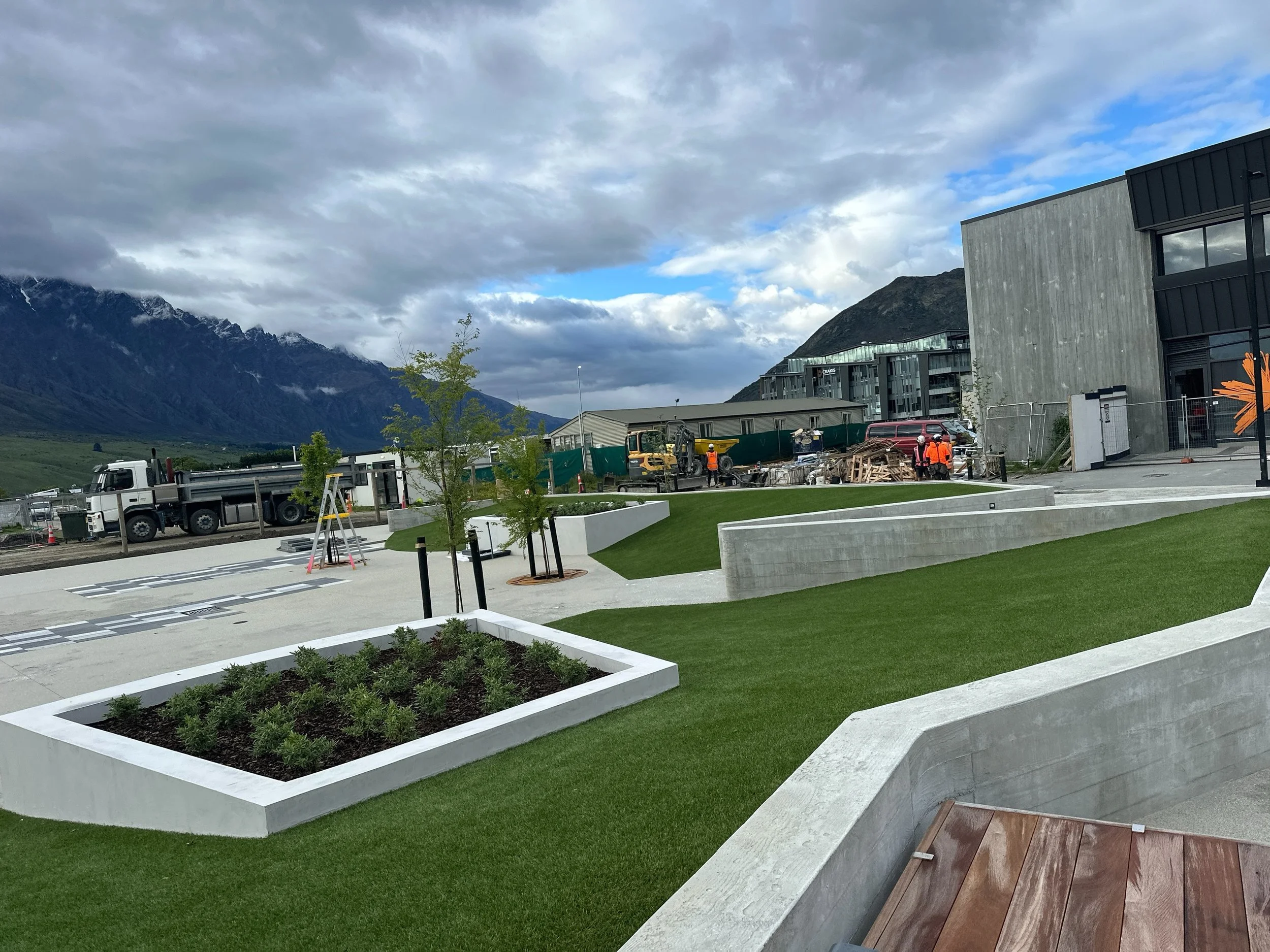 Modern landscaped development with construction workers and equipment beneath dramatic snow-capped mountains and cloudy skies