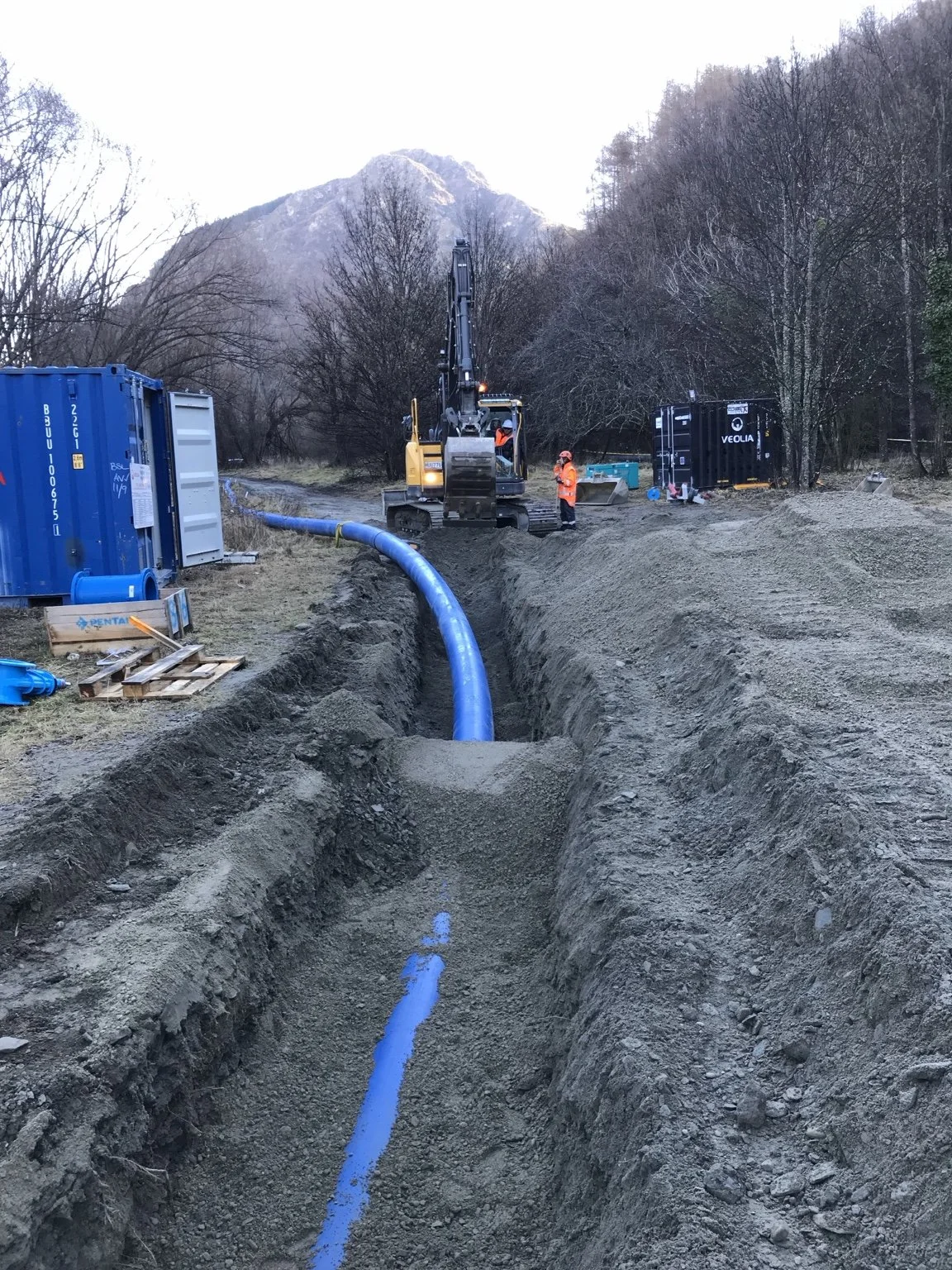 Excavator and workers installing a large blue pipe in a trench near a snow-dusted mountain