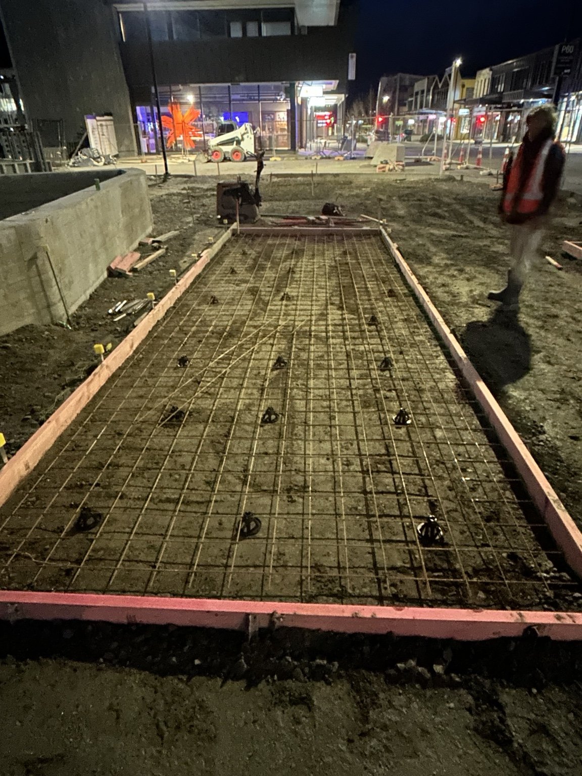 Nighttime construction site showing a rebar-reinforced concrete slab framed with wooden forms, a worker in a reflective vest, and a skid-steer loader.