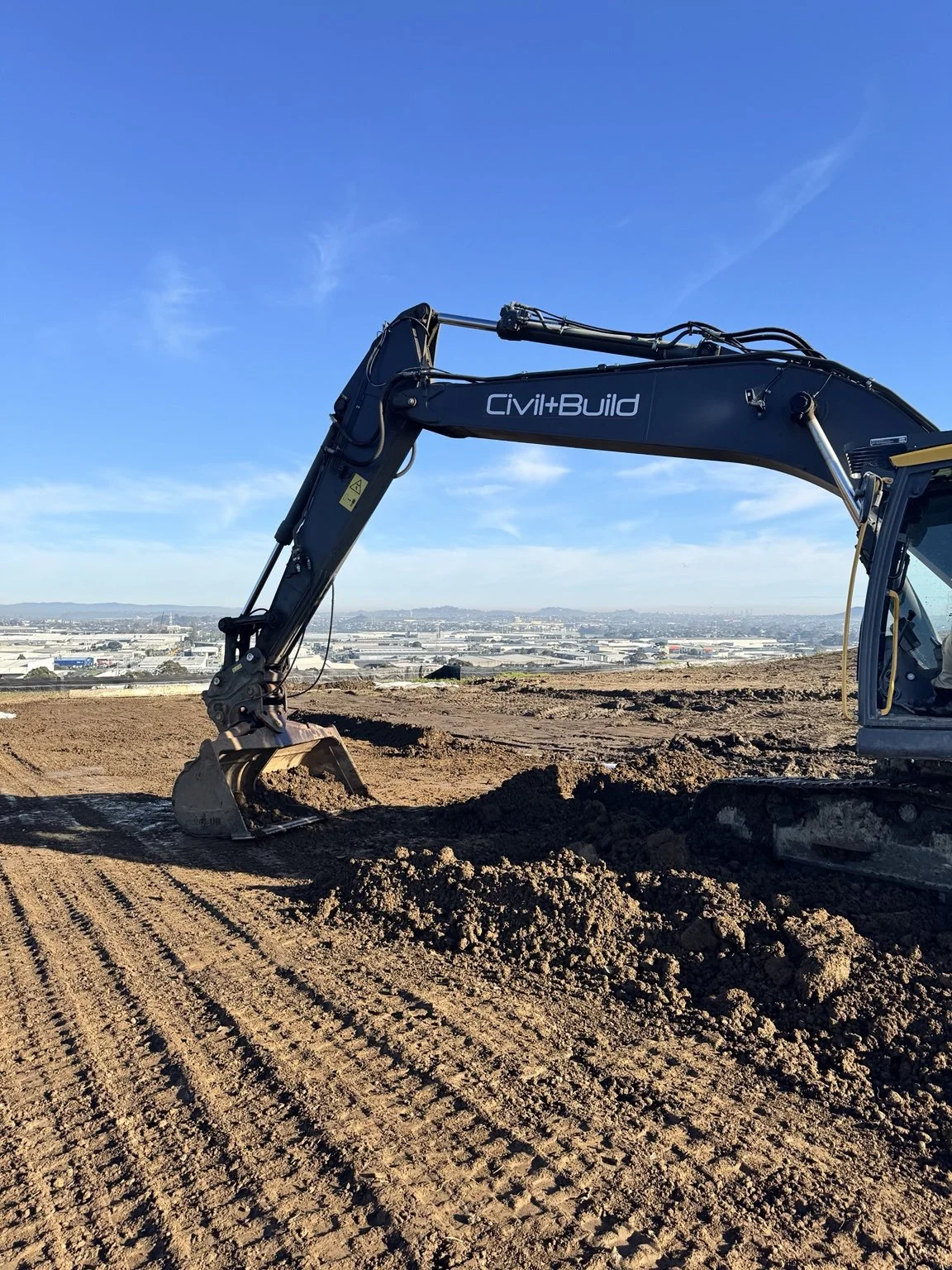 Excavator labeled “Civil+Build” digging at a dirt construction site with a cityscape in the background.