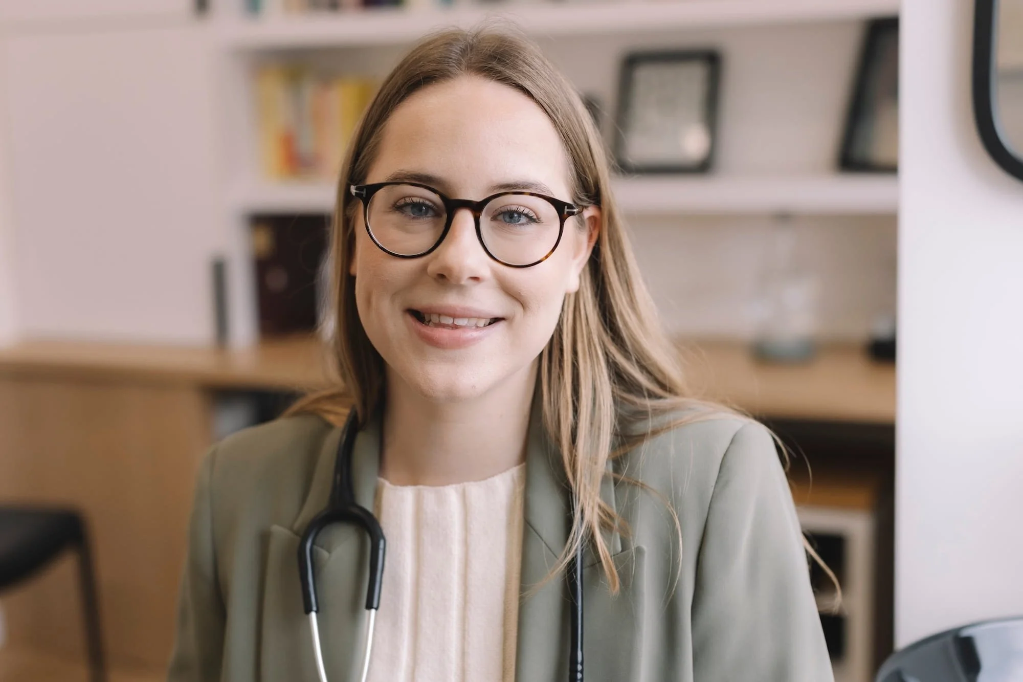 A smiling female healthcare professional wearing glasses and a stethoscope around her neck, working in a medical or office setting.