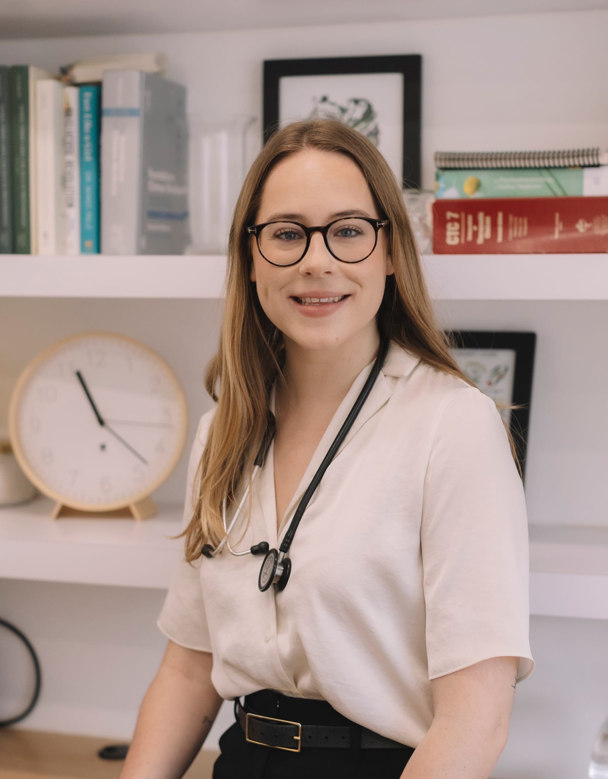 A woman with long reddish hair, wearing glasses, a beige top, and a stethoscope around her neck, standing in front of a shelf with books, a clock, and framed artwork.