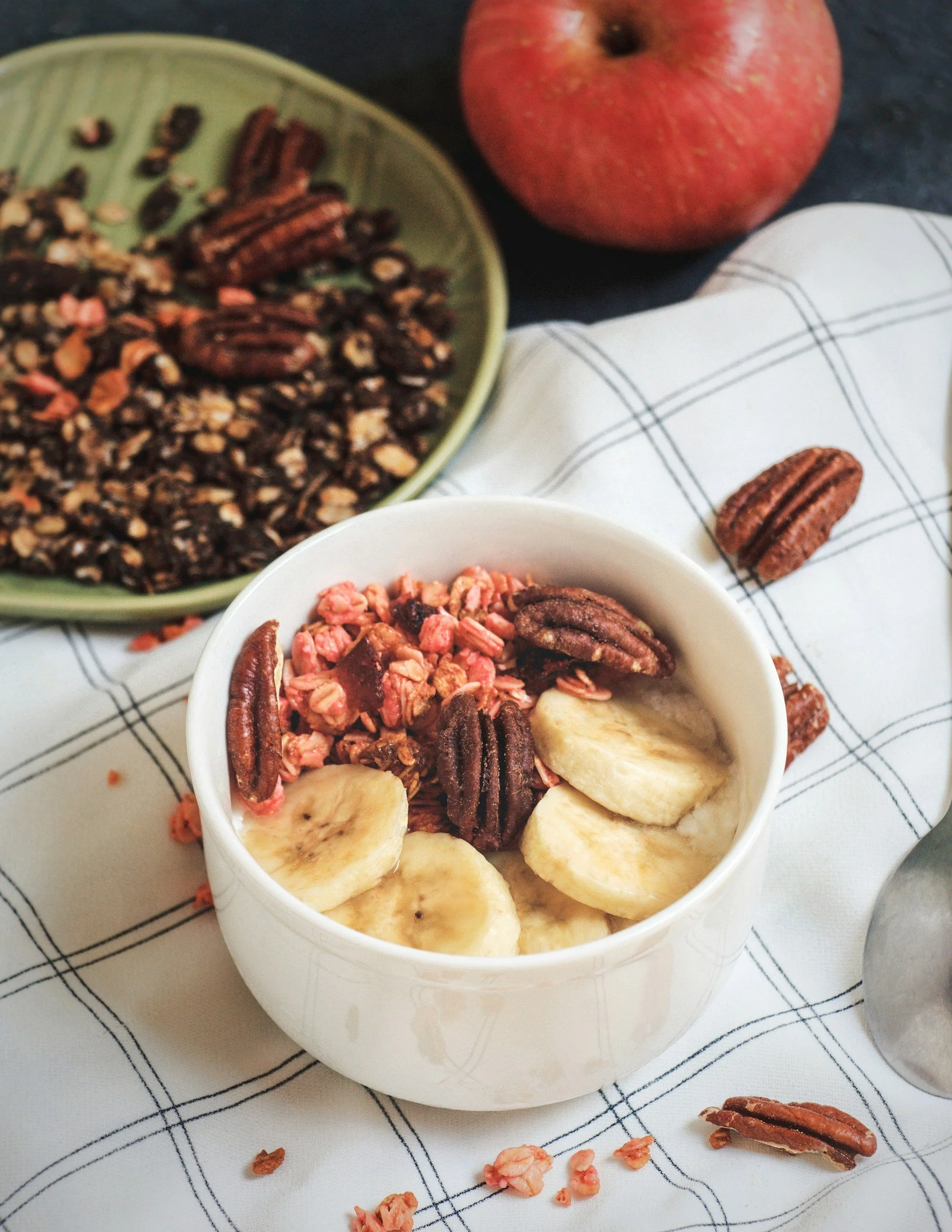 Close-up of a white bowl filled with granola, sliced bananas, and pecans on a black-and-white checkered cloth. Pecan pieces and granola are scattered around the bowl. A whole apple is partially visible in the background.