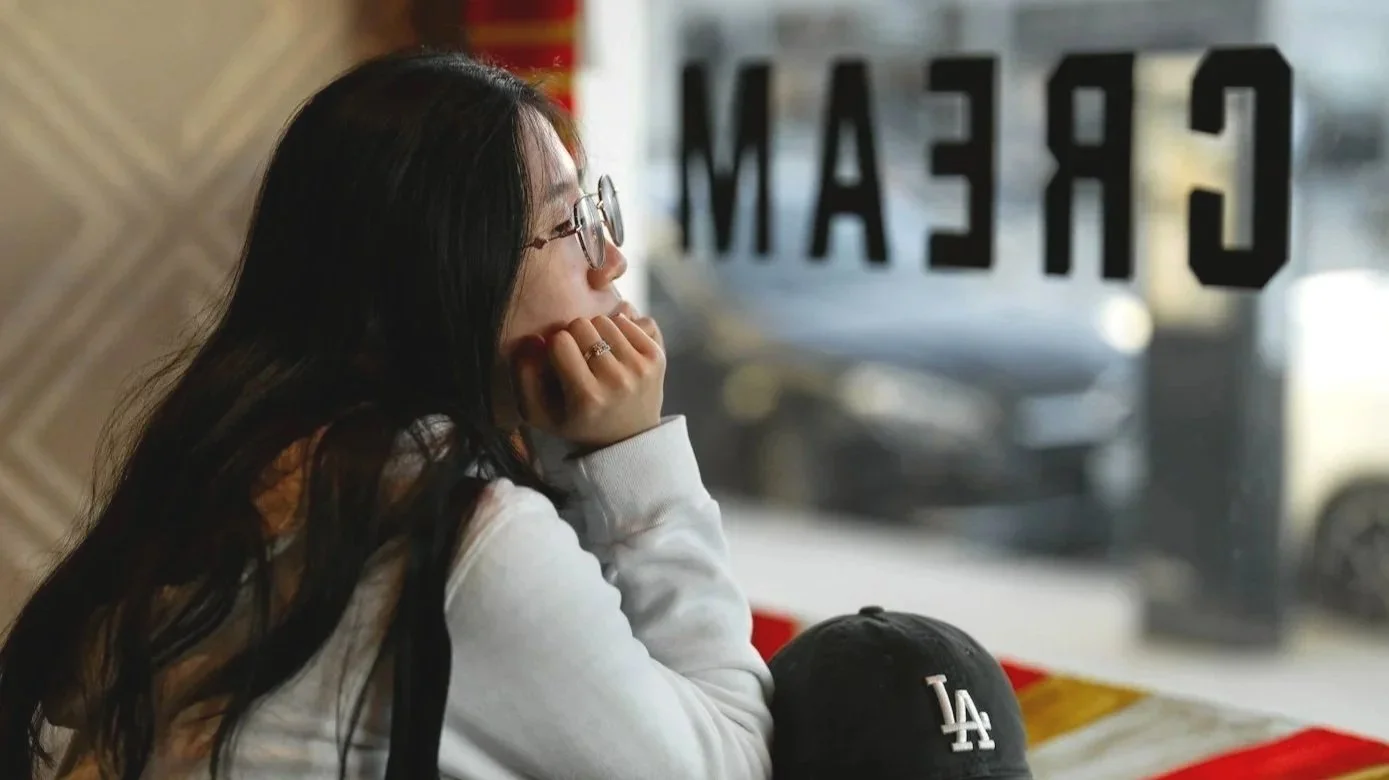 A woman with long dark hair wearing glasses and a white sweatshirt sitting inside a cafe, looking out the window with her chin resting on her hand. A black LA Dodgers cap is on the table in front of her. The window has 'CREAM' written on it in reverse, and a blurry street scene is visible outside.