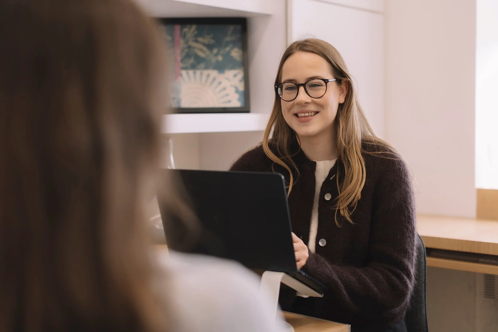 A woman with glasses and long hair smiling during a conversation in an indoor setting.