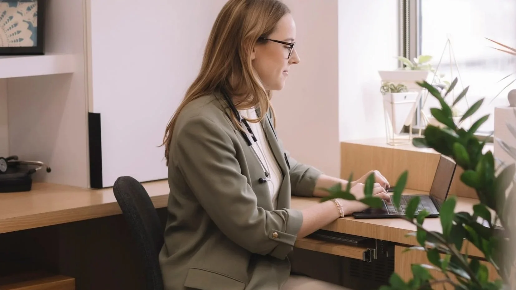 A woman with long red hair, glasses, and a stethoscope around her neck working on a laptop at a wooden desk in a bright office with plants by the window.
