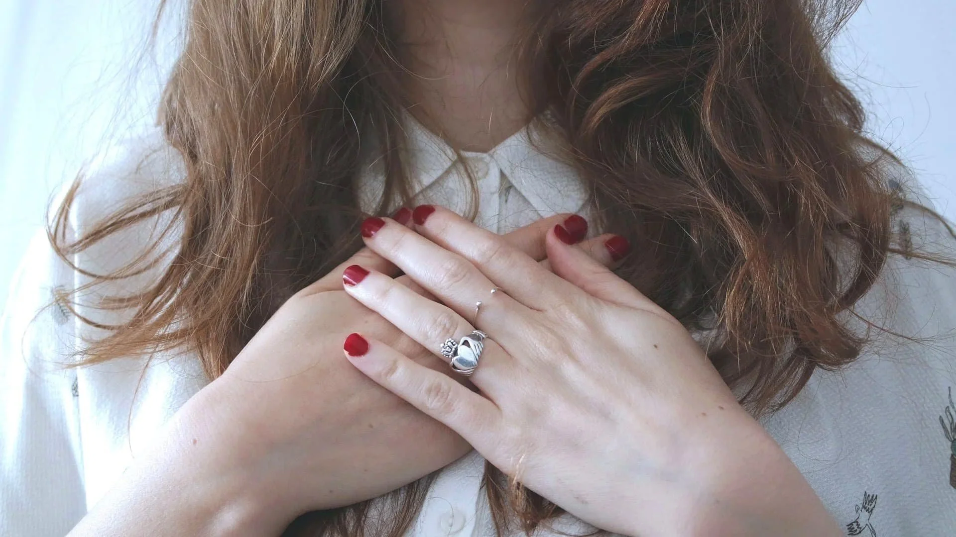 A woman with wavy red hair wearing a white shirt, with her hands crossed over her chest, displaying rings on her fingers and red nail polish.