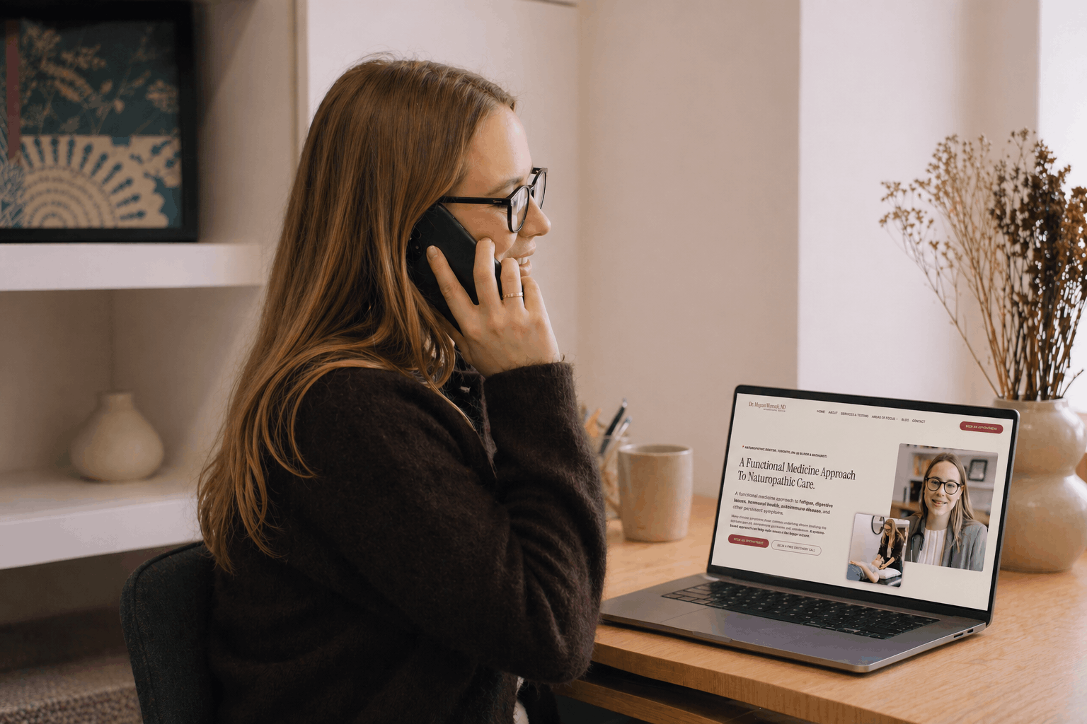 Woman with glasses on a video call using a phone at her desk, with a laptop displaying a website about naturopathic medicine.
