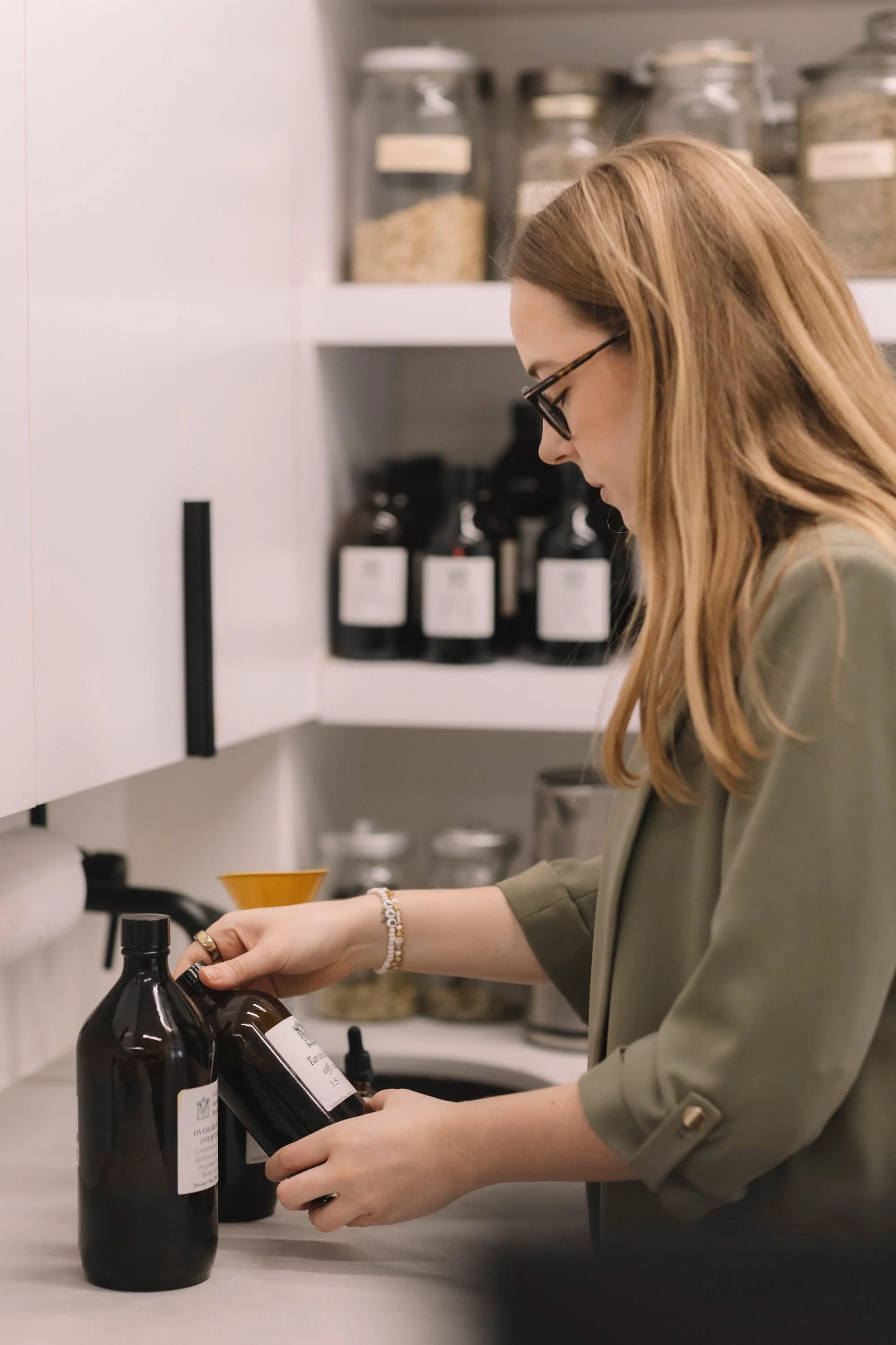 A woman with long, light brown hair, wearing glasses and a greenish-gray jacket, is organizing dark bottles with white labels in a kitchen or lab setting. Behind her on the shelves are glass jars and black bottles.