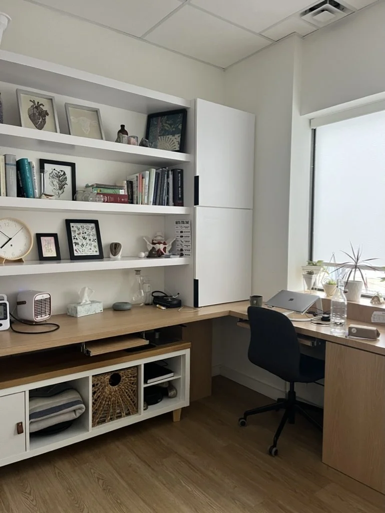 A home office with a wooden desk, black office chair, and white shelves filled with books, framed pictures, and decorative objects. There's a window with potted plants on the windowsill, and various items like a laptop, water bottle, tissue box, and other office supplies on the desk.