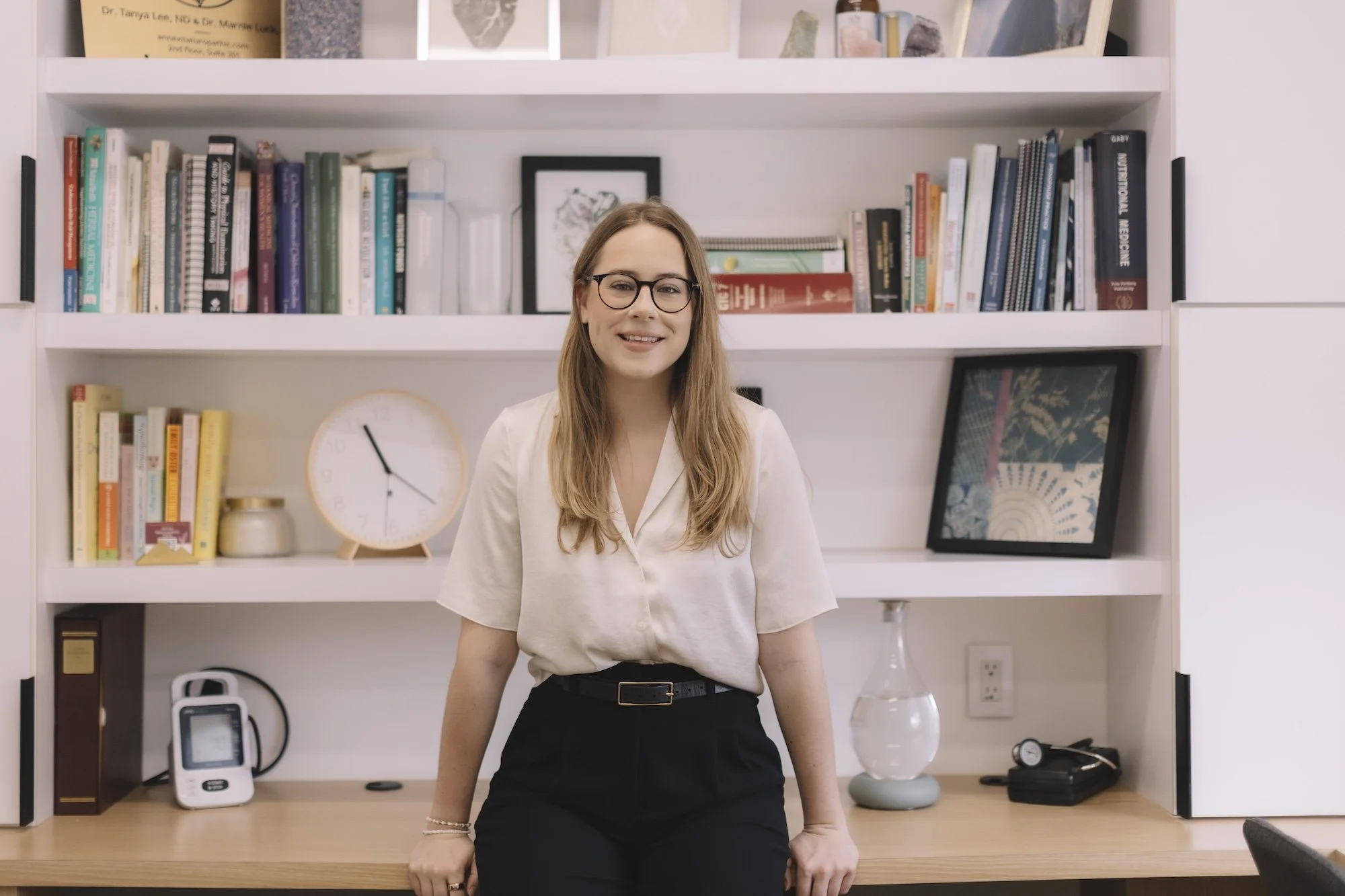 A woman with glasses and long hair, wearing a white blouse and black pants, standing in front of a bookshelf with books, framed pictures, and decorative items, smiling at the camera.