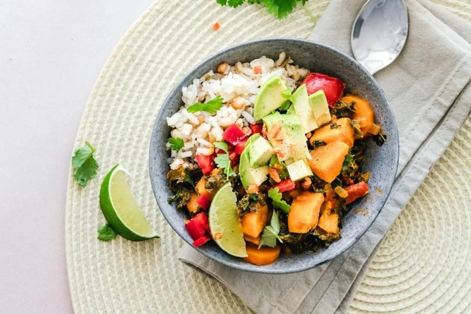 A bowl of rice and vegetable stir-fry topped with avocado, red peppers, and cilantro, served with lime wedges on the side.