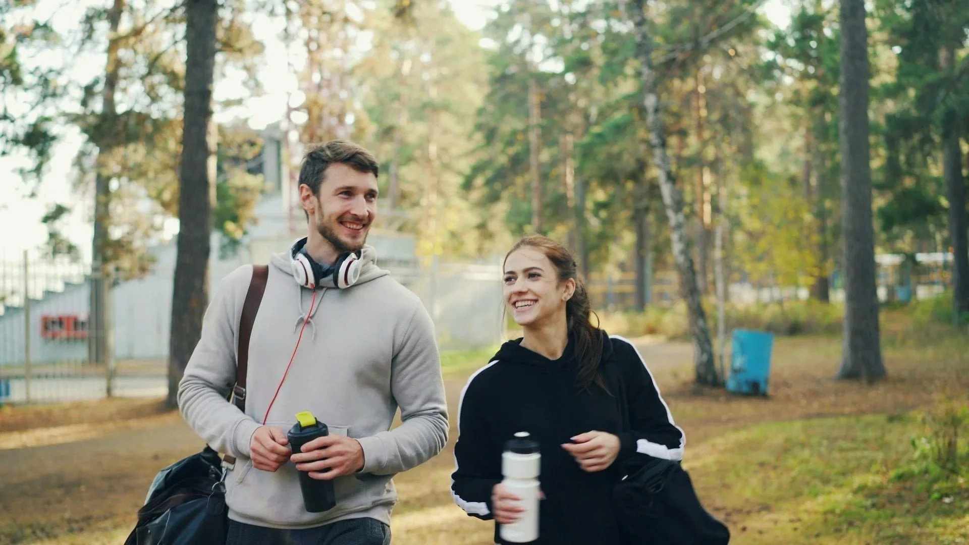 A young man and woman jogging together outdoors in a park during autumn, smiling and carrying water bottles.