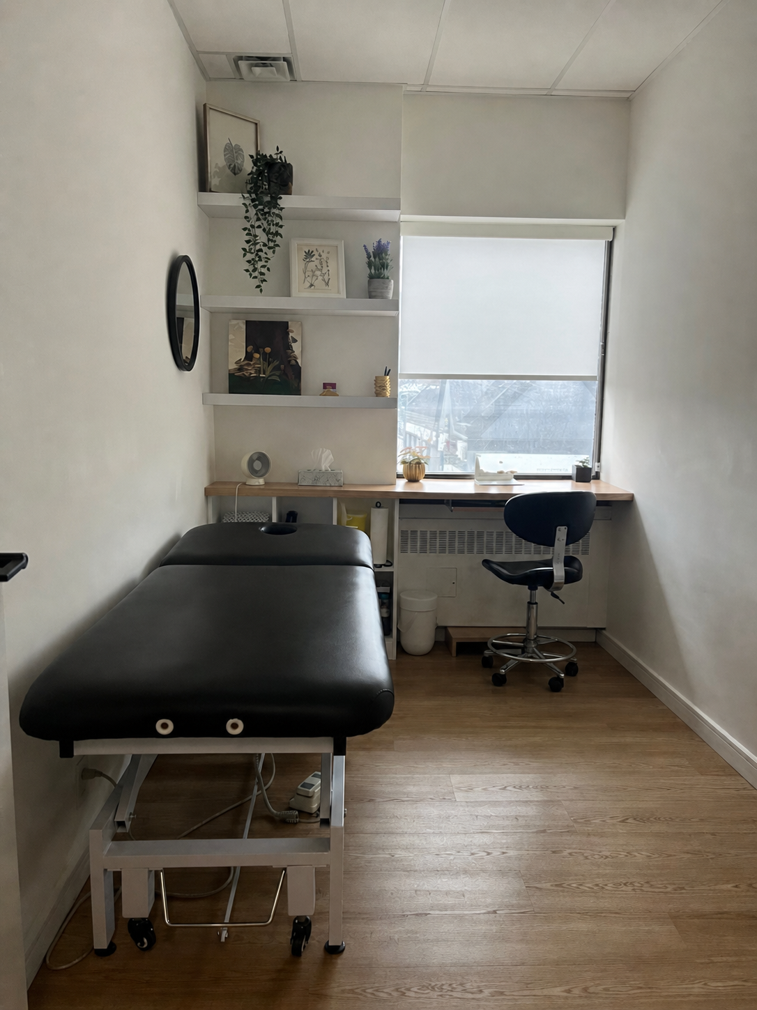 A small room designed for medical consultations with a bed on the left, a desk with a black office chair near the window, shelves with decorative items, and a window with a roller shade.