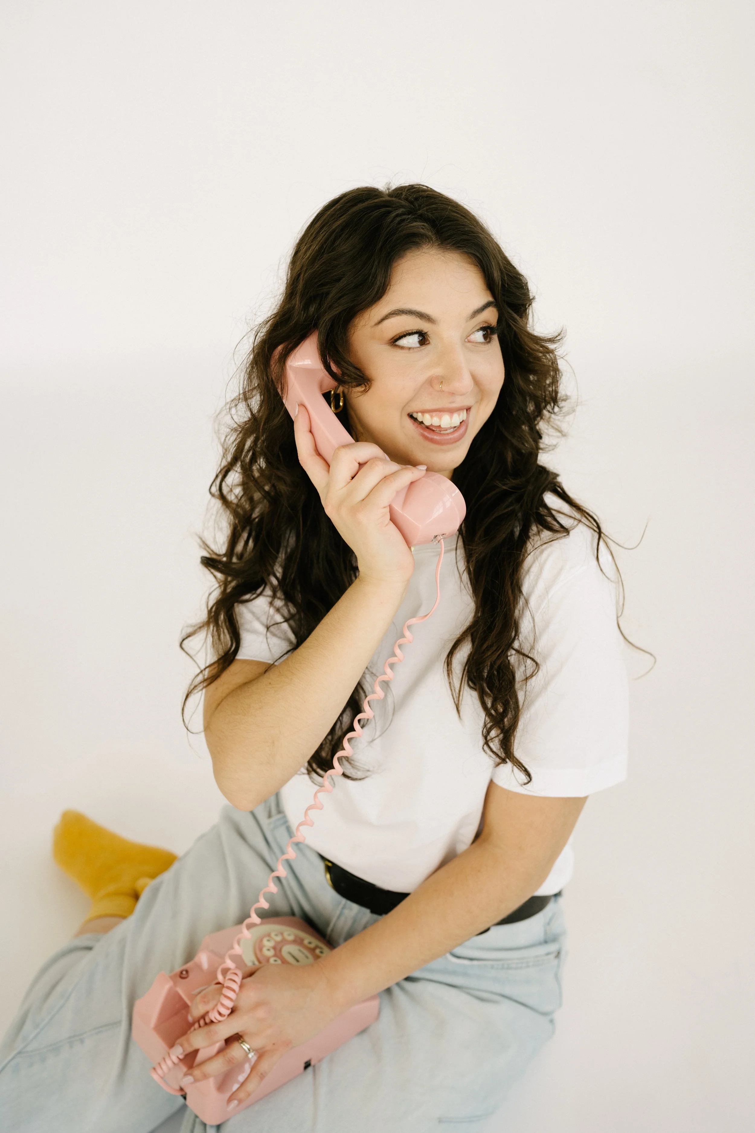 A young woman with dark, curly hair smiling and talking on a vintage pink rotary phone, sitting on the floor against a plain white background, wearing a white t-shirt and light jeans.