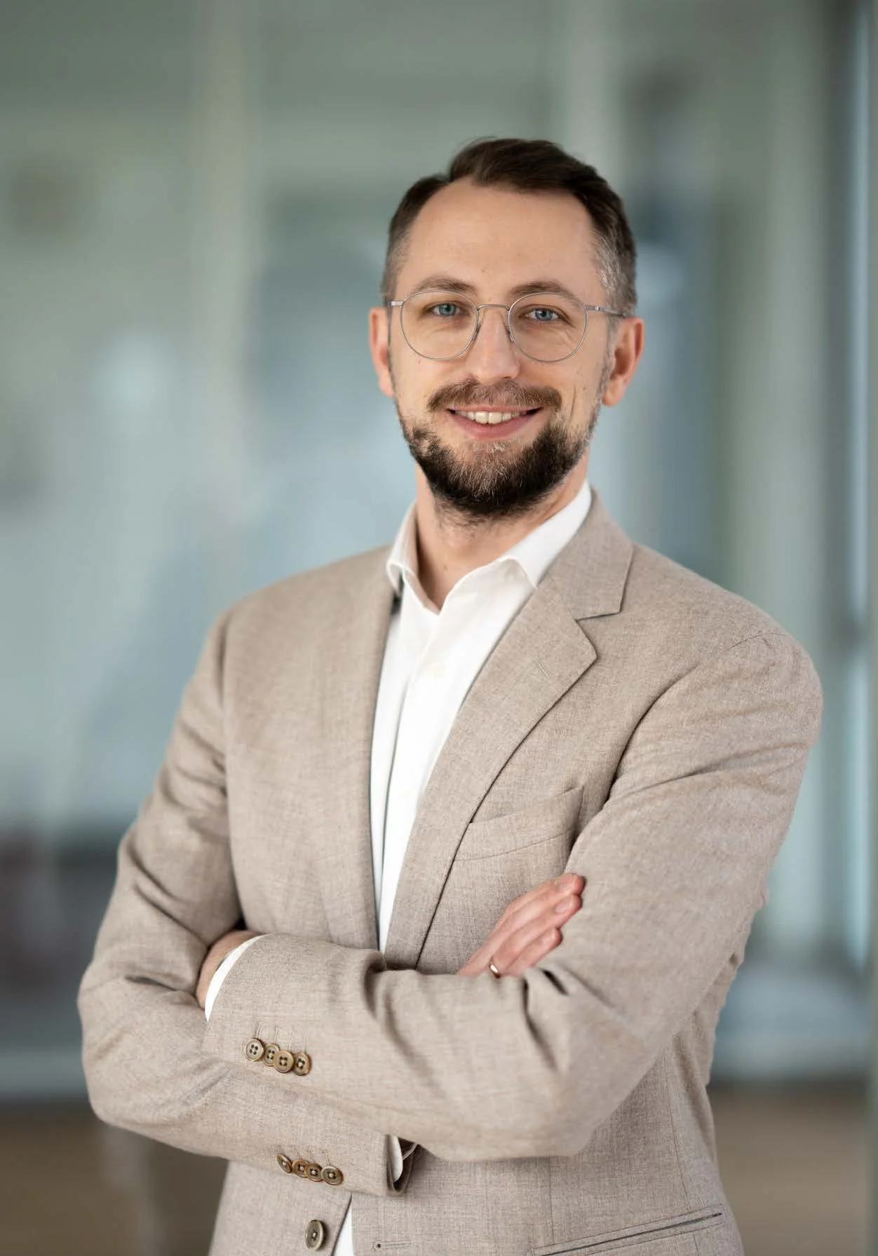 Ein Mann in einem beigefarbigen Anzug und weißem Hemd, lächelnd mit verschränkten Armen vor einem Bürohintergrund.