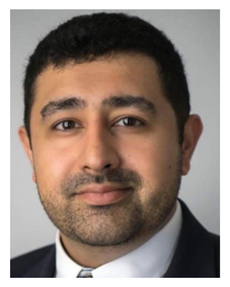 A professional headshot of a man with short black hair, brown eyes, and a beard, wearing a suit and tie against a plain gray background.