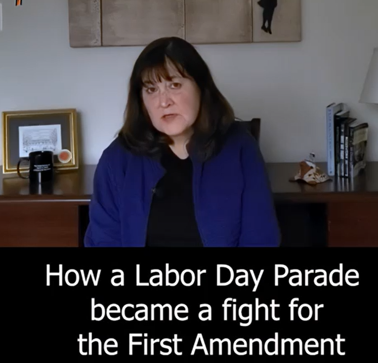 Woman with dark hair and glasses sitting in front of a desk, wearing a blue jacket, with text overlay about a Labor Day Parade and the First Amendment.