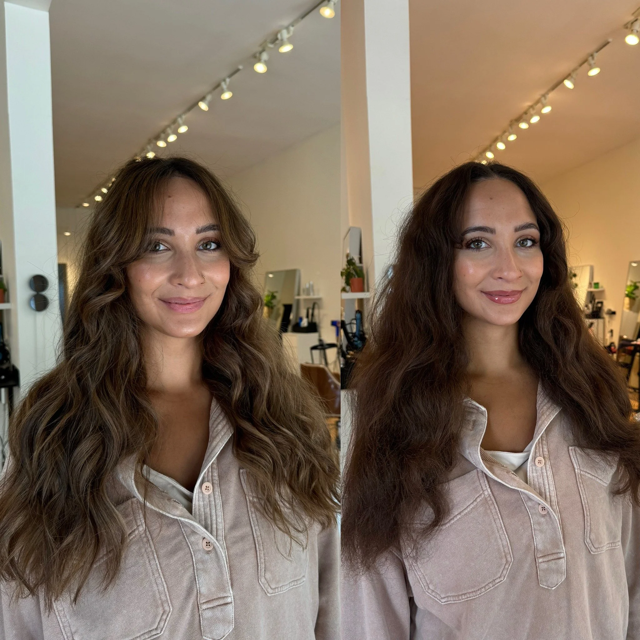 Two women with long, wavy brown hair and light skin, smiling in a salon or studio with hair styling equipment and plants in the background, wearing light-colored shirts with open collars.