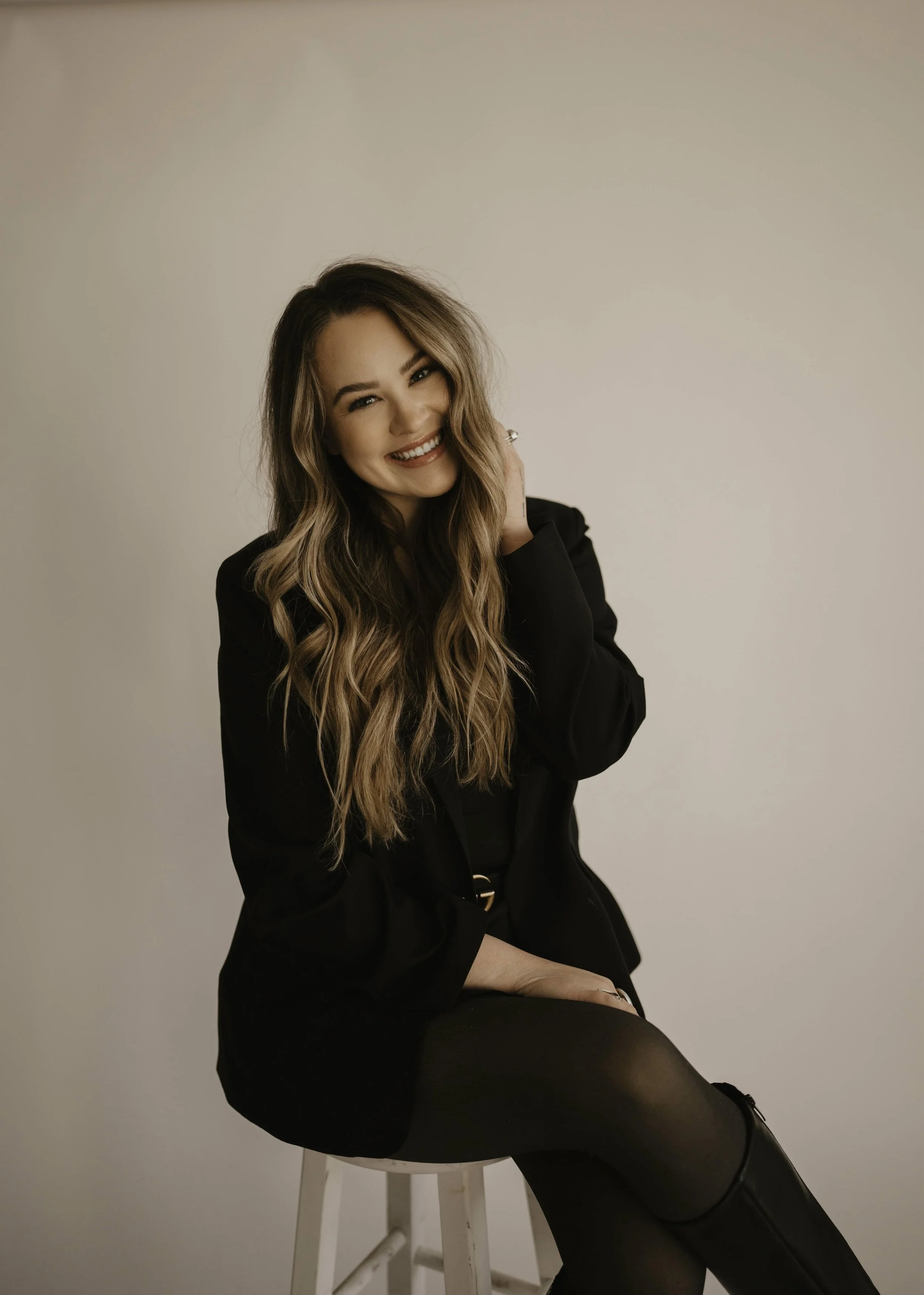 A young woman with long wavy hair, smiling and winking, sitting on a white stool against a plain beige wall. She is dressed in black clothing, including a blazer, tights, and pants.swilly hair co