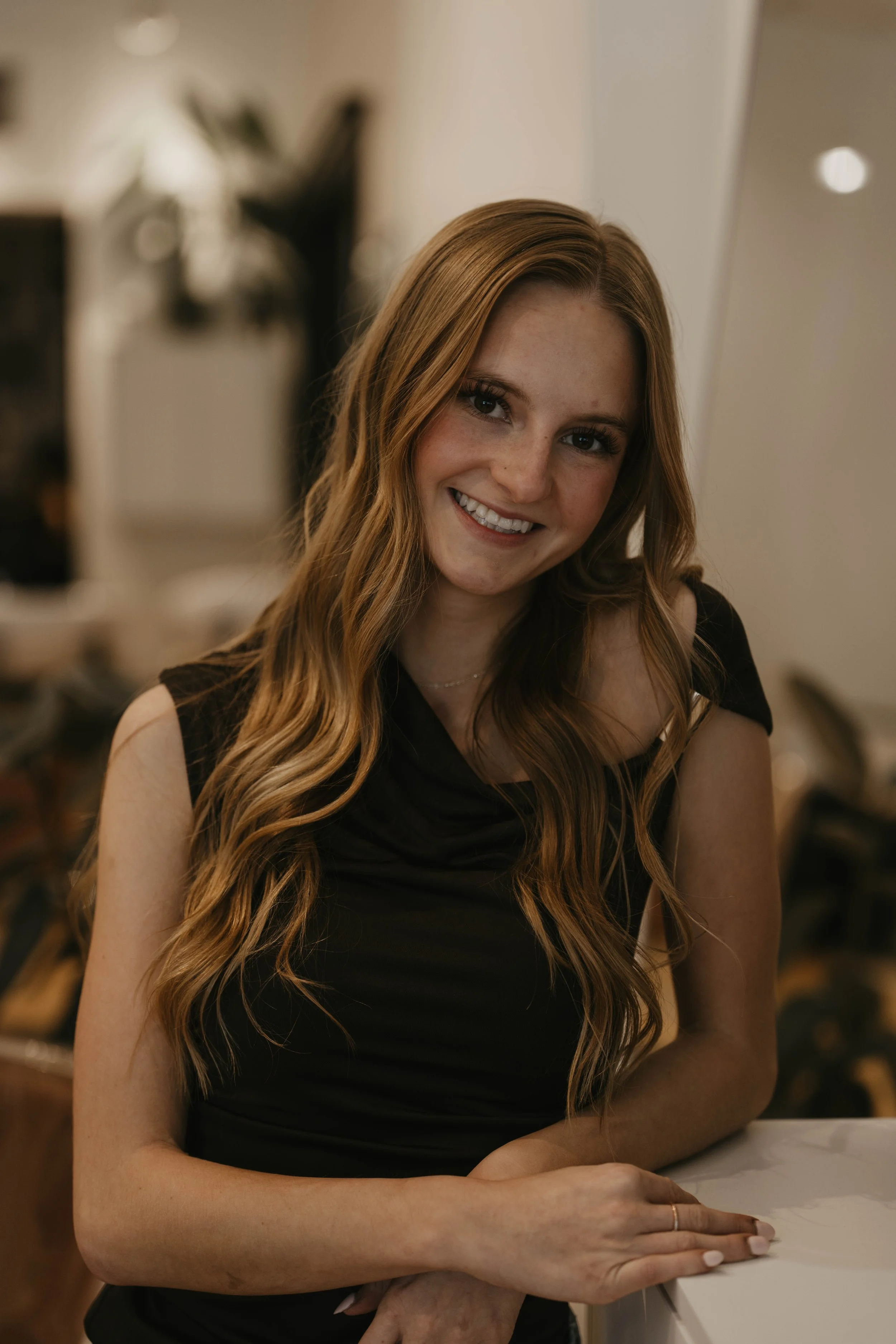 A smiling woman with long wavy red hair in a black sleeveless top sitting at a white table indoors.