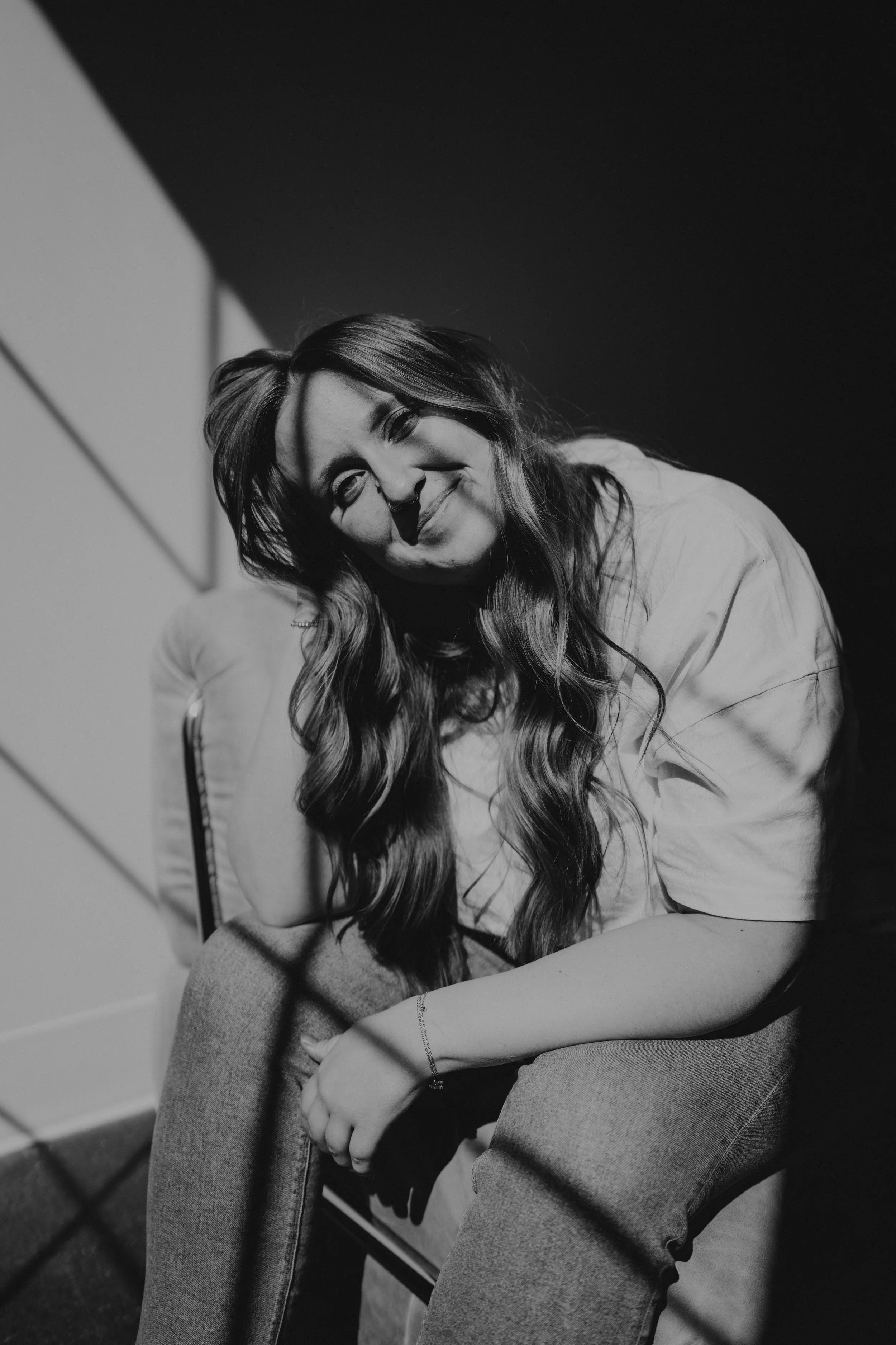 A black and white photo of a young woman with long, wavy hair, smiling gently. She is seated, with her arms resting on her lap, and is wearing a light-colored shirt and jeans.