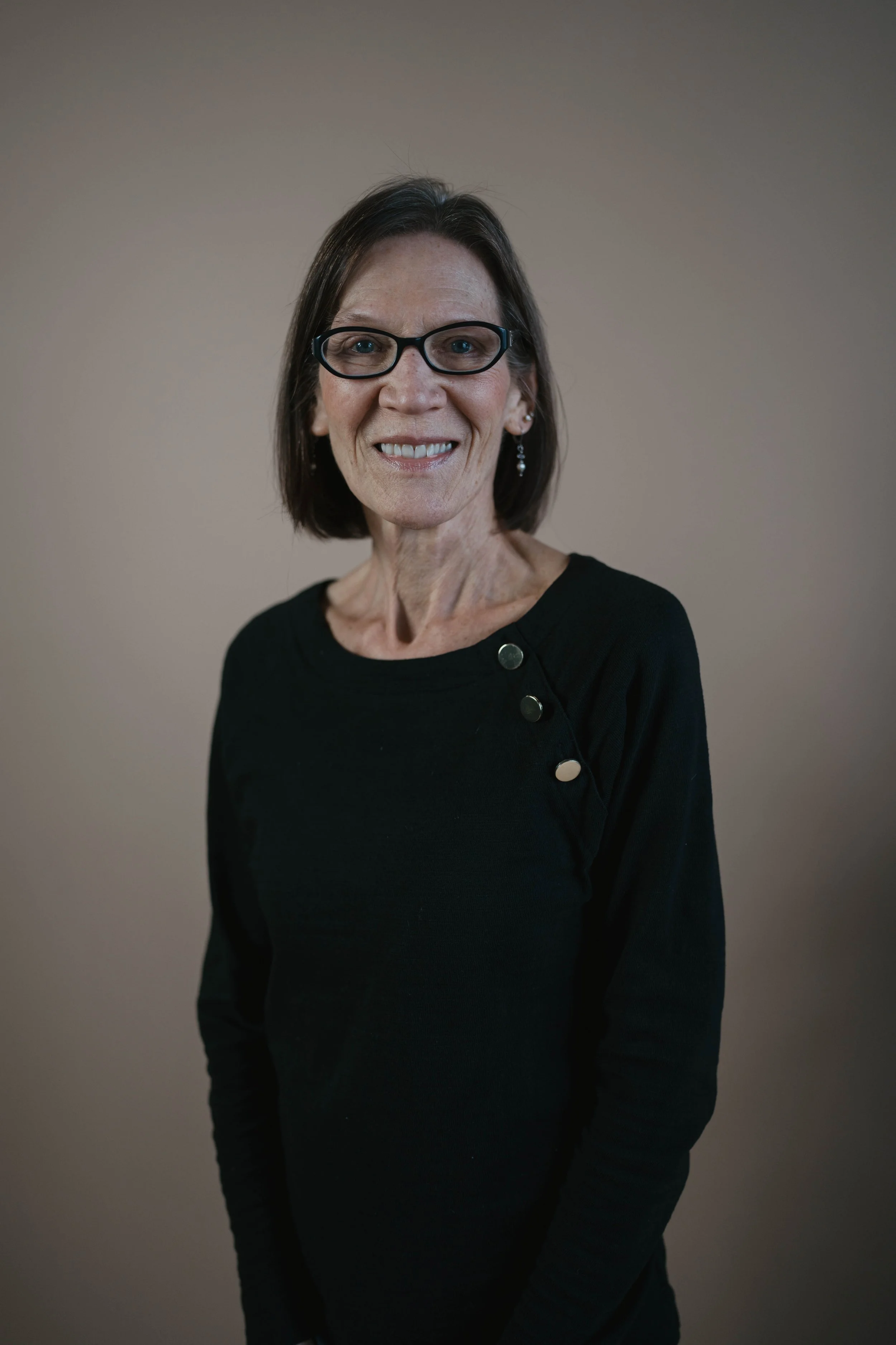 A middle-aged woman with short dark hair, wearing glasses and a black top with metallic buttons on the shoulder, smiling at the camera against a plain beige background.