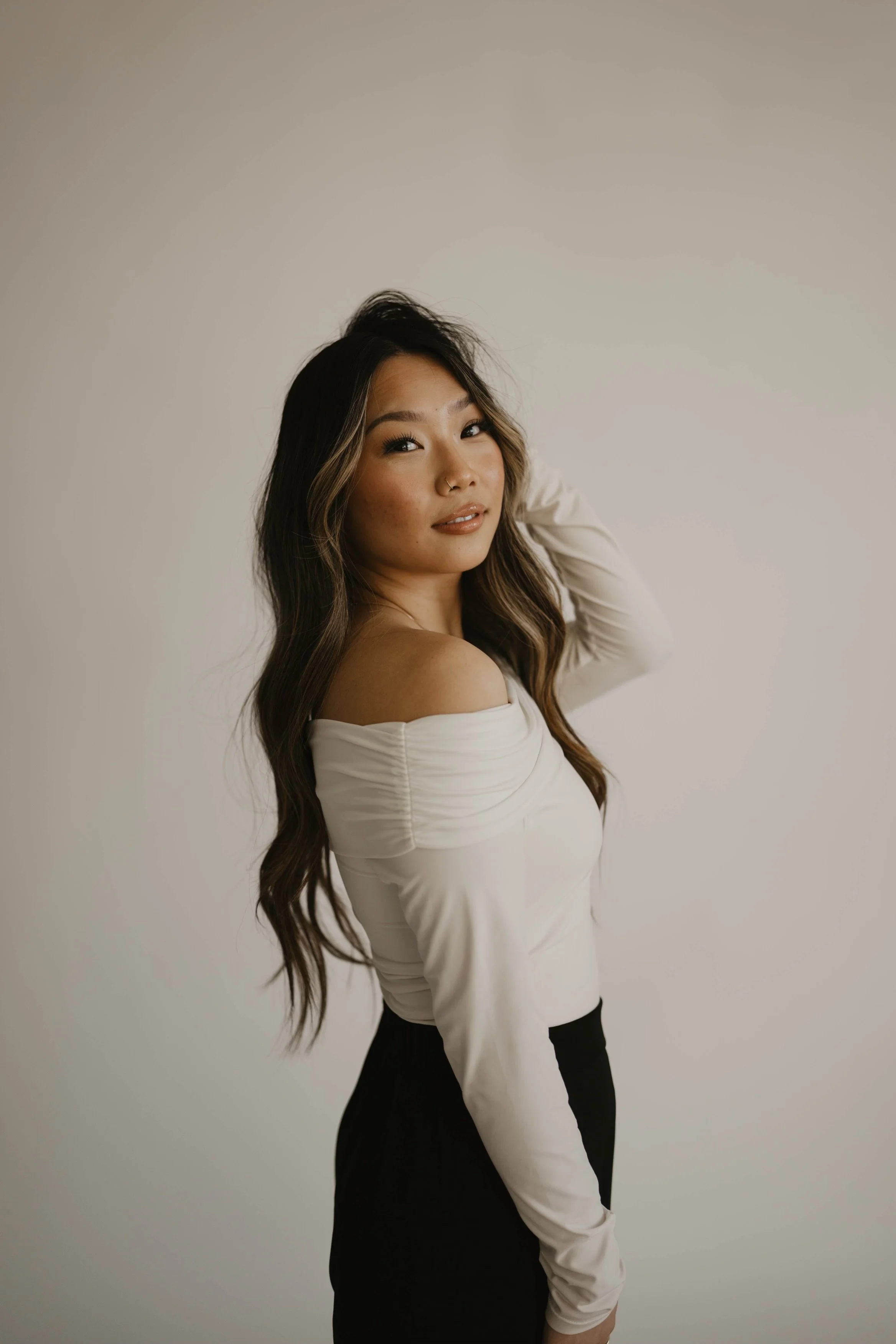 A young woman with long wavy hair, wearing a white off-the-shoulder top and black pants, stands against a plain white background. She has a slight smile and is looking at the camera with her hand resting on her head.
