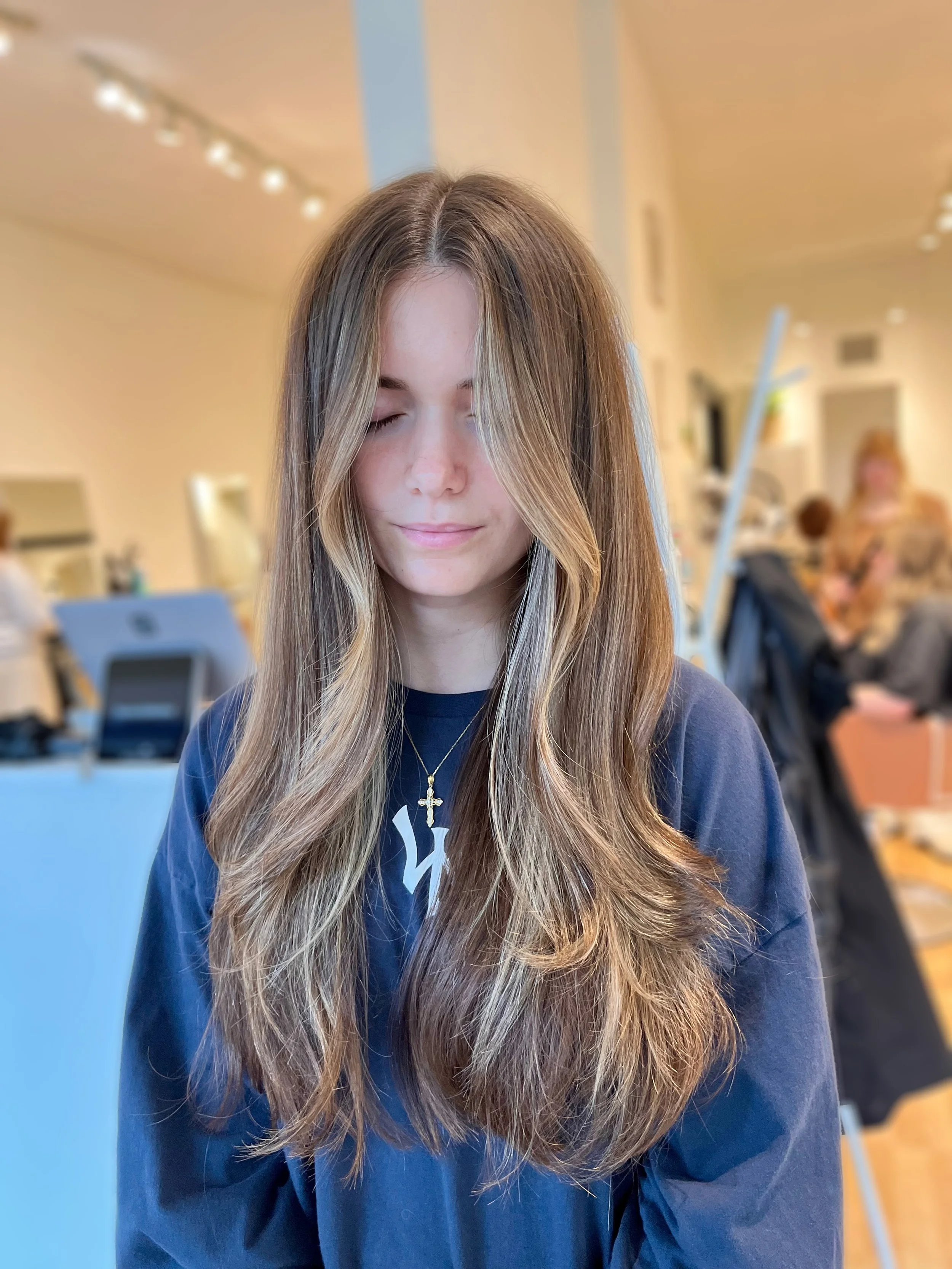 A young woman with long, wavy brown hair and light skin, wearing a blue shirt and a gold cross necklace, eyes closed, in an indoor setting that appears to be a hair salon or beauty shop with blurred background of other people and salon equipment.