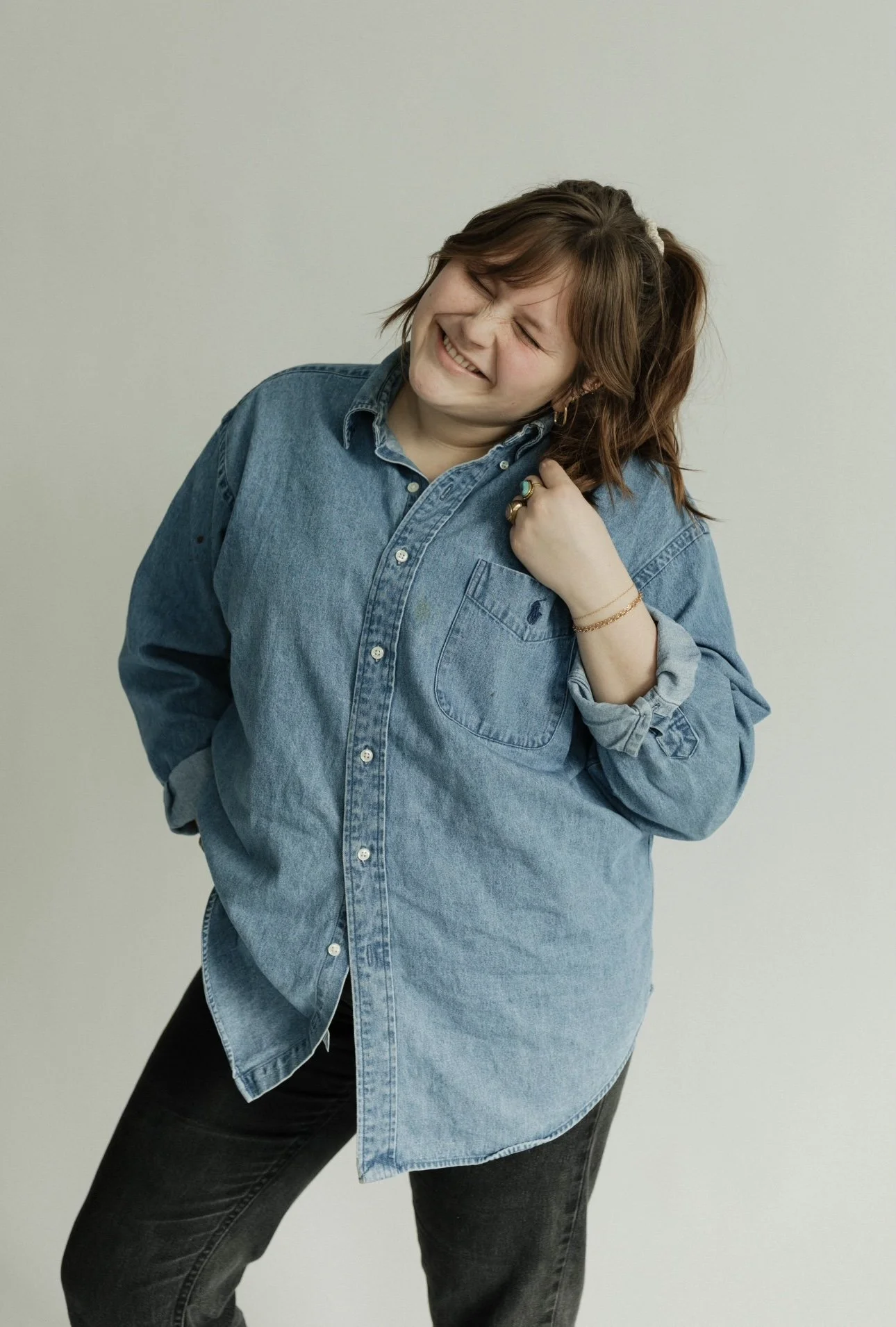 Woman in a denim shirt and black pants smiling with eyes closed, standing against a plain light background. Maija