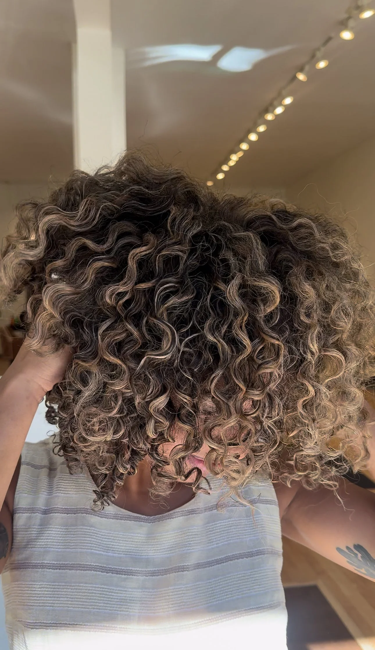 Person with short, curly textured hair that is dark brown with blonde highlights, wearing a striped shirt, in a salon with track lighting.