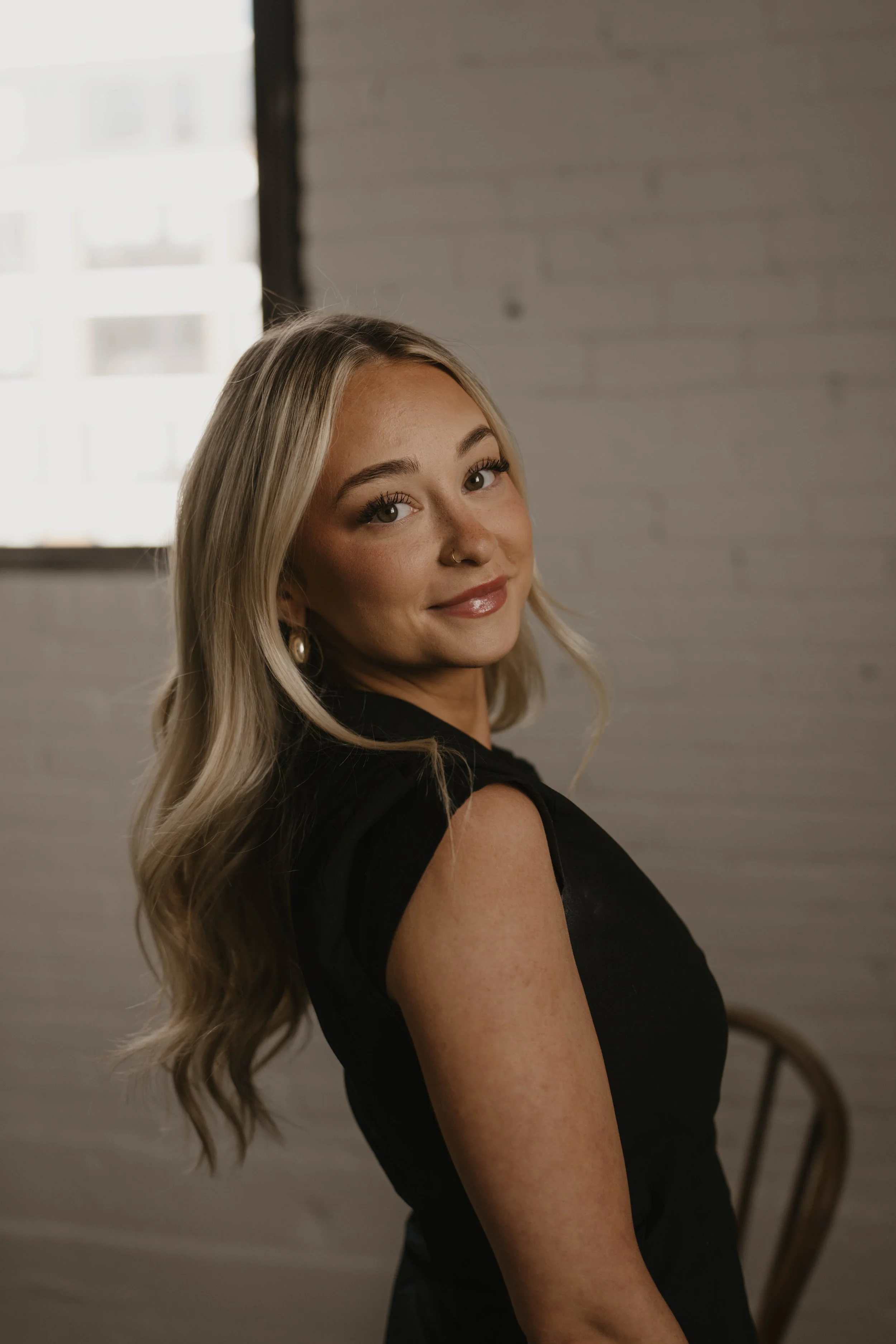 A young woman with long, wavy blonde hair smiling and looking over her shoulder, wearing a black sleeveless top, with a wooden chair nearby and a white brick wall in the background.
