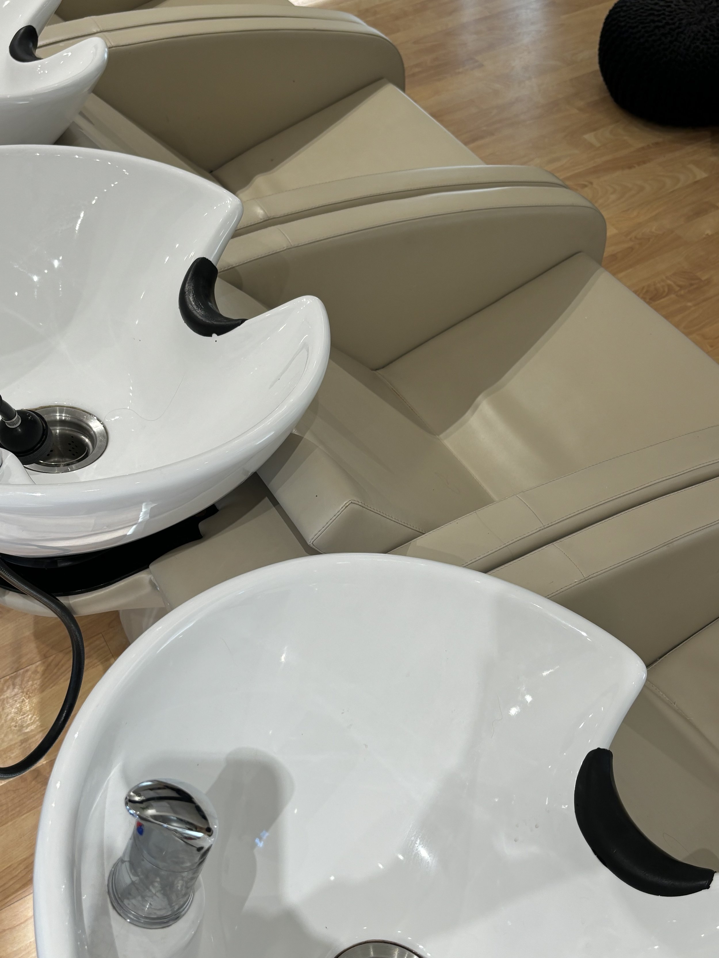 Close-up of a white salon sink in a hair salon, with a chrome faucet and a black spray nozzle, surrounded by beige seating and a wooden floor.